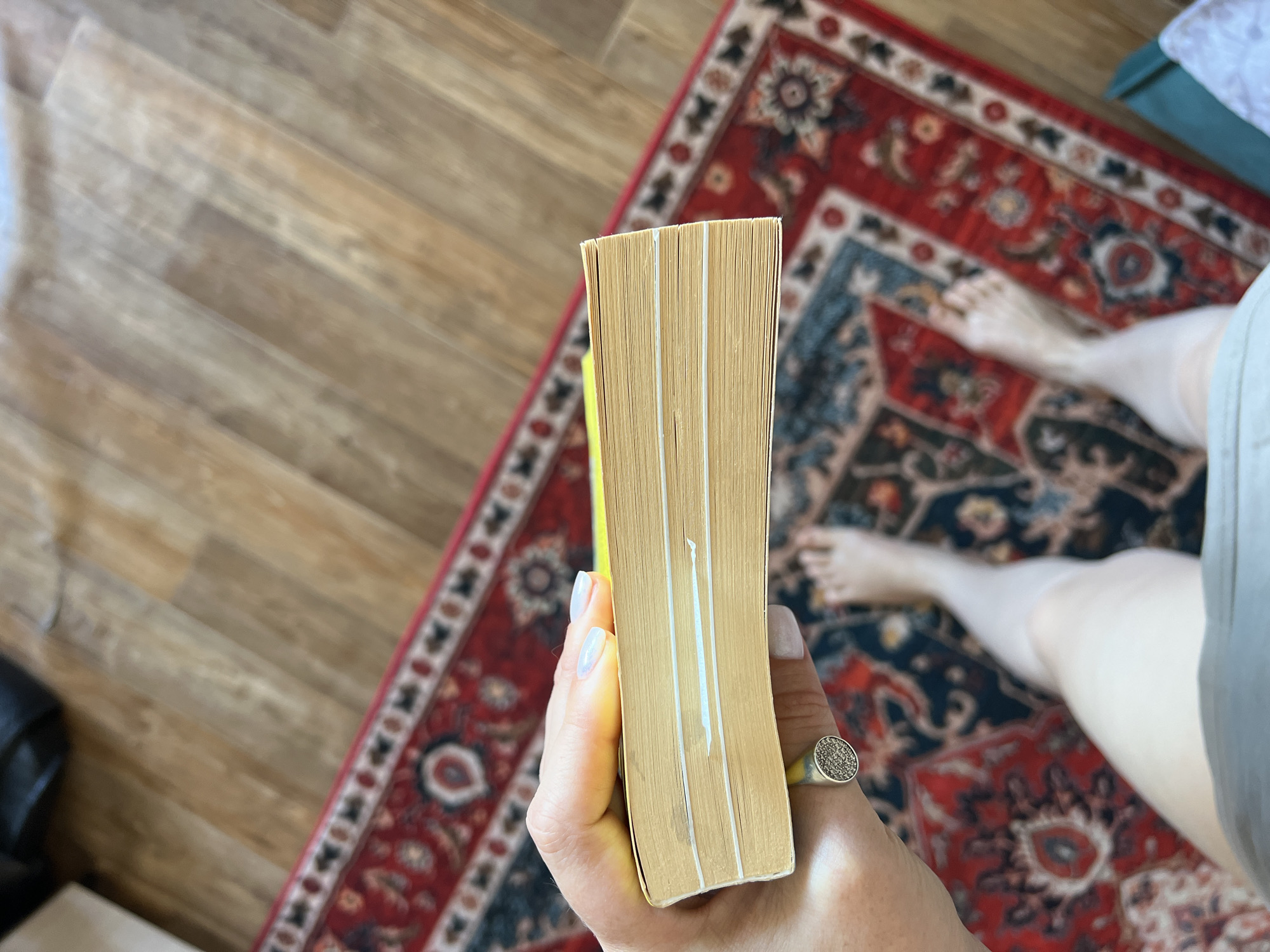 The image shows a close-up view of a hand holding a thick yellow book. The book's pages are visible, and there appear to be several white elastic bands wrapped around it. The hand has light-colored nail polish and is wearing a ring with small, dark stones. In the background, a person's legs and feet are visible, along with a red and blue patterned rug and wooden flooring. The lighting suggests an indoor setting. The specific location in Novosibirsk, Russia cannot be determined from the provided image.