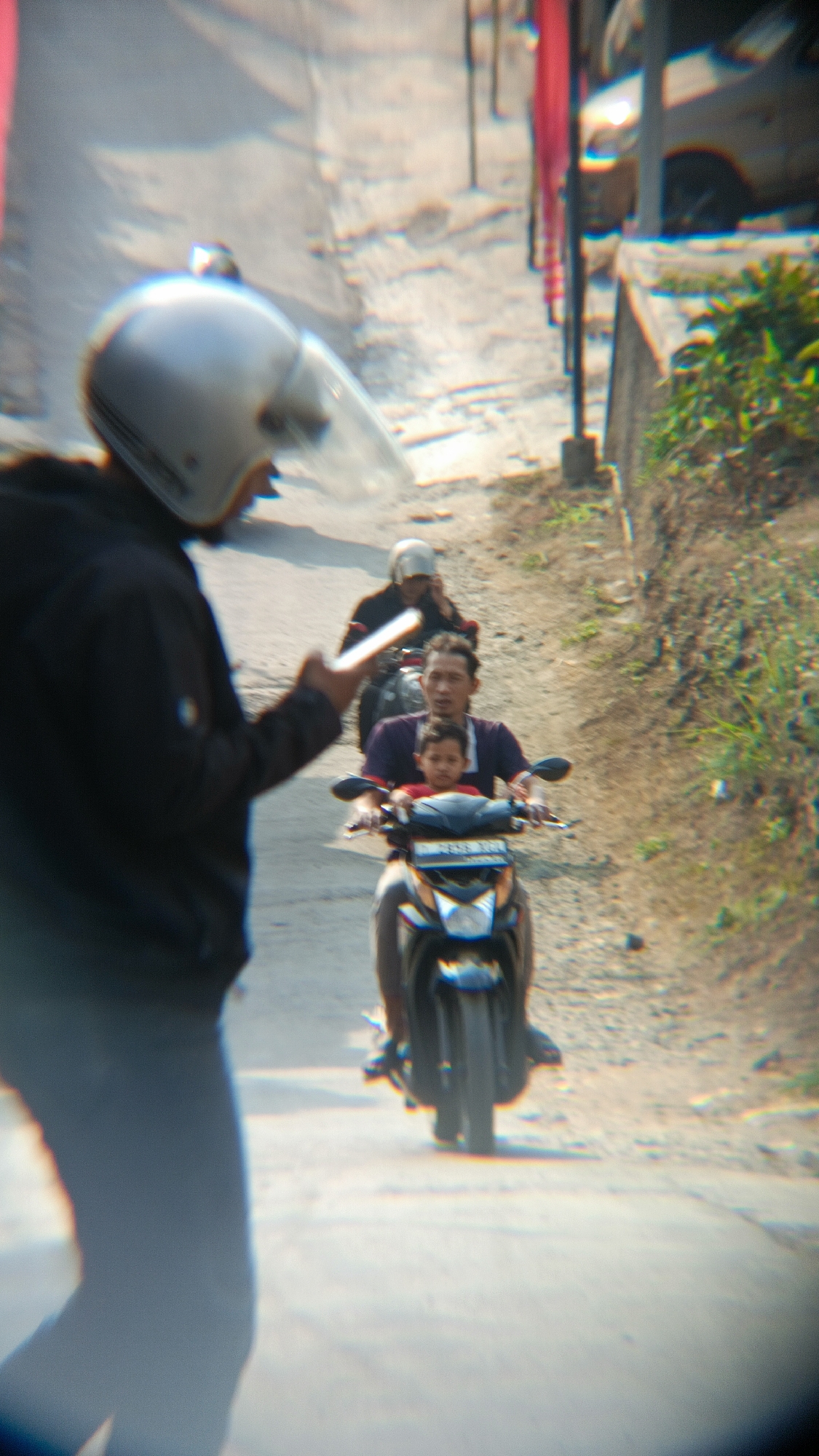 In Margajaya, Indonesia, a person wearing a silver helmet and a black jacket is captured from behind, walking on a dirt road. In front of them, two motorcycles are visible. The first motorcycle carries two individuals: a man wearing a purple shirt and a child wearing a red shirt. The second motorcycle is further ahead, with a person wearing a light-colored helmet. The road is dusty and uneven, with vegetation visible on the right side of the frame. The overall scene depicts a casual moment of travel on a rural road.