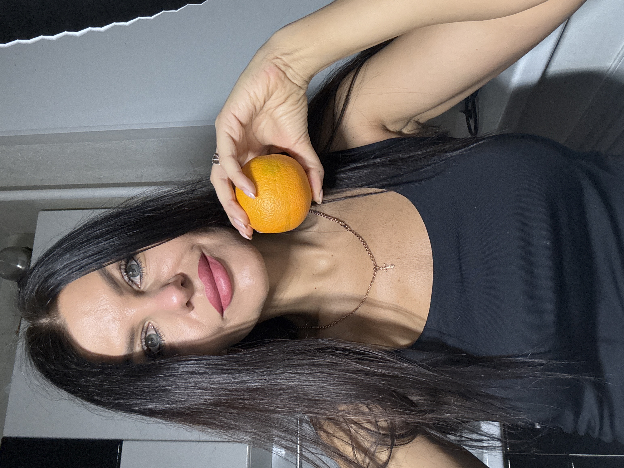 A sideways-oriented, medium close-up shot features a woman posing indoors, holding an orange. The woman has long, straight, dark brown hair that frames her face and cascades over her shoulders. She has fair skin, pronounced eyebrows, light-colored eyes (likely blue or grey), and wears pink lipstick. She is wearing a black sleeveless top or dress and a delicate rose gold-toned chain necklace with a small cross pendant visible near her collarbone. A ring is visible on her right hand, which holds the orange.
Her right arm is bent, and she is holding a small, ripe orange, which appears to be positioned near her shoulder or cheek. She looks directly towards the camera with a neutral to slightly smiling expression. The background suggests an indoor setting with a light grey or white wall/ceiling area and some darker, possibly tiled, elements below. The lighting appears to be artificial, casting some soft shadows. There is no visible text. The context for this image is Chaikovsky, Russia.