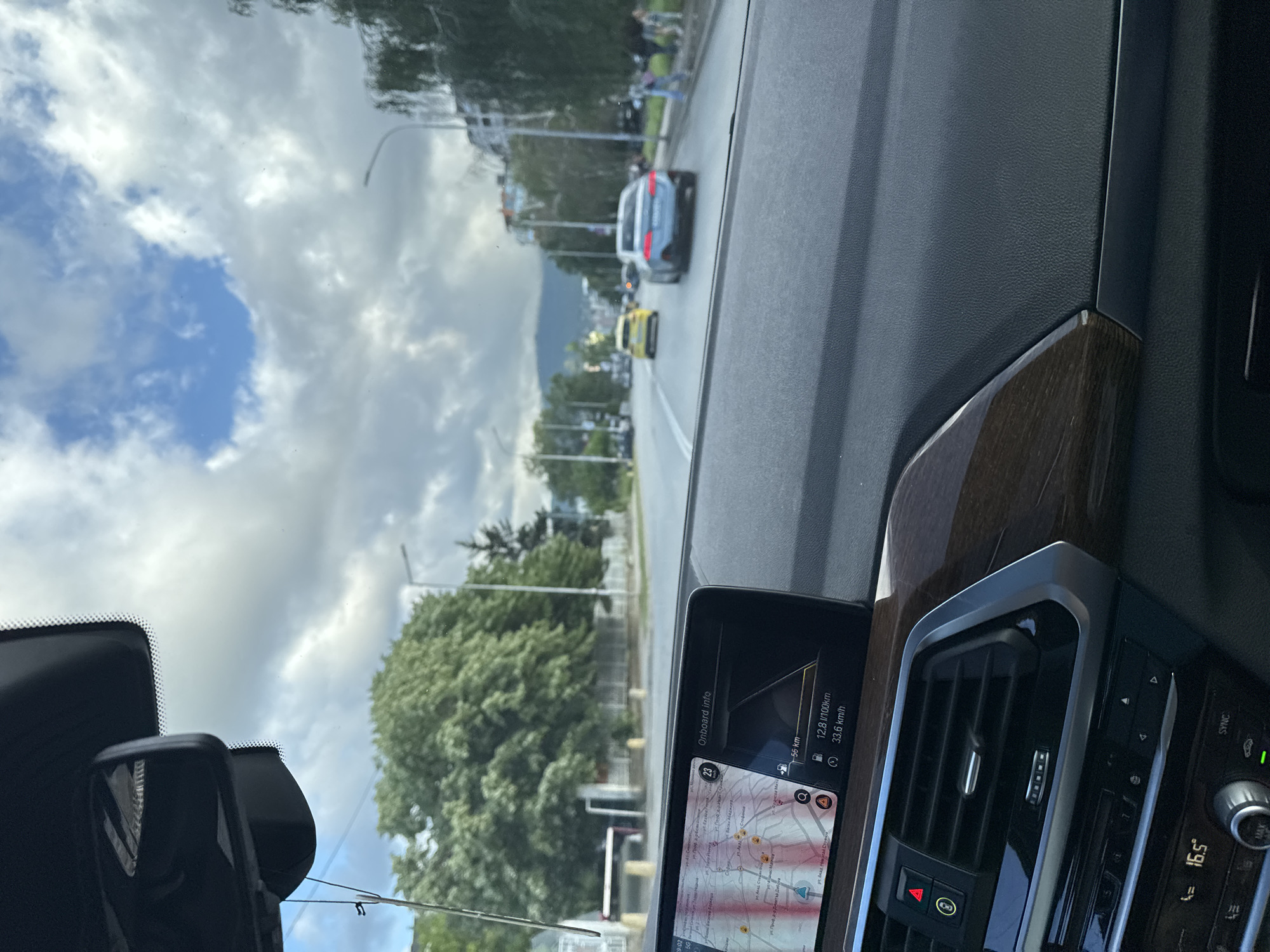 The image provides a driver's perspective from inside a vehicle, looking out onto a street in Sofia, Bulgaria. The car's interior features a dashboard with a prominent infotainment screen and wood-grain trim on the passenger side. A rearview mirror assembly is partially visible at the top left of the frame.
The infotainment screen displays navigation information, including a map with street names written in Cyrillic, such as "ул. Акад. Борис Стефанов," confirming the location context. Onboard data indicates a remaining distance of "56 km," fuel consumption of "12.8 l/100km," and a current or average speed of "33.6 km/h." The ambient temperature reads "16.5°." Other visible text on the screen includes "Onboard info," "Q" (likely for search), and "A" (a car icon). The top-left corner of the image shows "9:02" and "5G," likely from a phone's status bar reflected on the windshield or dashboard. A "SYNC" button is visible on the center console next to air vents and controls.
Outside the car, a multi-lane road is visible with other vehicles, including a silver SUV and a yellow car, suggesting moderate traffic. Lush green trees line the street, and buildings and distant mountains are present in the background. The sky is partly cloudy, with areas of bright blue contrasting with white clouds, indicating daytime. The overall impression is a daytime drive through an urban environment with natural elements.