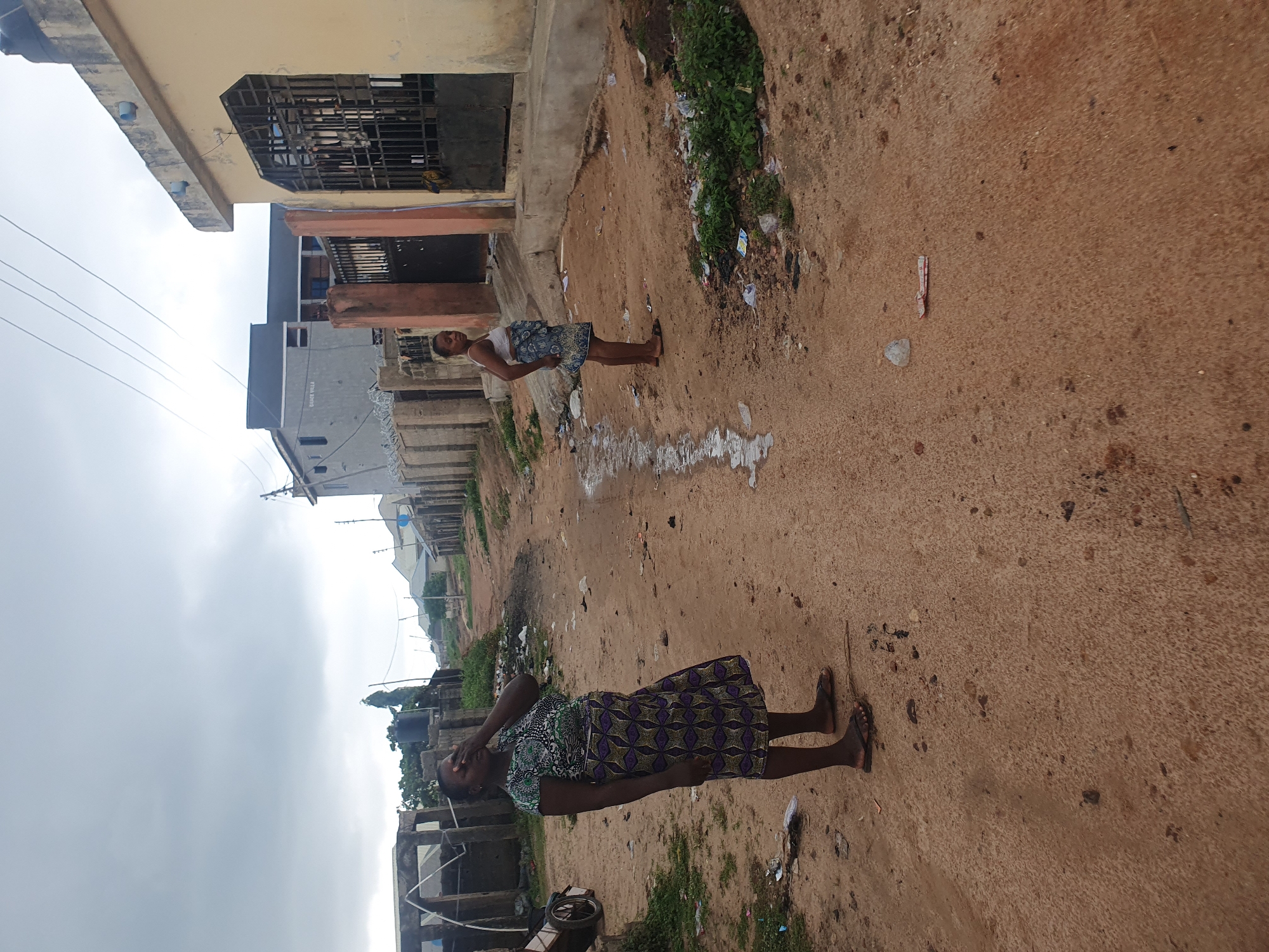 In Lokoja, Nigeria, on a dirt road in front of buildings, a woman stands with her right hand covering her face. She is wearing a patterned dress and sandals. To her right, a younger woman in a patterned skirt and top stands with her hands clasped in front of her. The road is muddy with puddles and scattered debris. Behind the women, buildings of varying architectural styles line the street, some with barred windows. The sky is overcast.