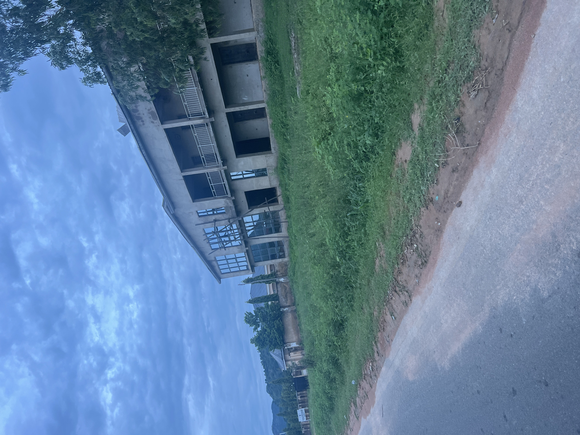 The image shows a building under construction in Kona Garu, Nigeria. The building is multi-story with concrete walls and visible balconies. The windows are large and reflective, some appearing to be glass panes and others are empty openings. The building is surrounded by lush green grass and a dirt path, with a paved road running alongside. Trees and foliage are visible in the foreground and background, suggesting a rural or semi-urban setting. The sky is overcast with clouds, indicating a potentially gloomy or late afternoon time of day. There are no people visible in the image. The overall impression is one of an incomplete structure in a natural landscape.