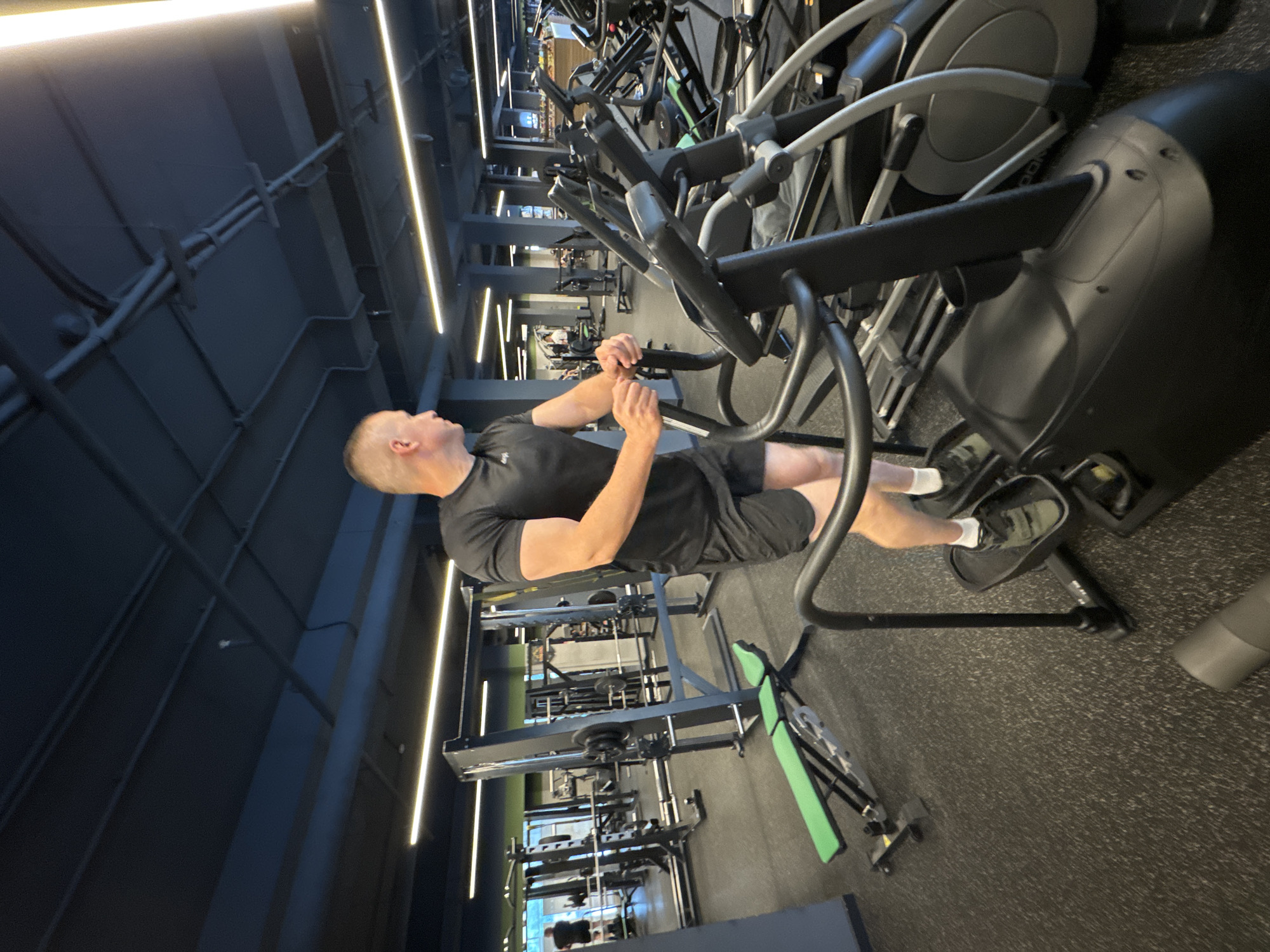 A man with a shaved head, wearing a black t-shirt and black shorts with white socks and athletic shoes, is exercising on an elliptical machine in a gym. The gym appears to be modern with a dark gray floor, black gym equipment, and linear fluorescent lighting fixtures on the ceiling. The man is in profile, facing left, with his hands on the handlebars of the elliptical, and his feet on the pedals. His expression is not visible. In the background, other gym equipment and possibly other people can be seen, though they are out of focus. The lighting suggests it could be daytime or evening, as the gym is well-lit. The overall scene conveys a sense of a typical workout session in a well-equipped fitness facility.