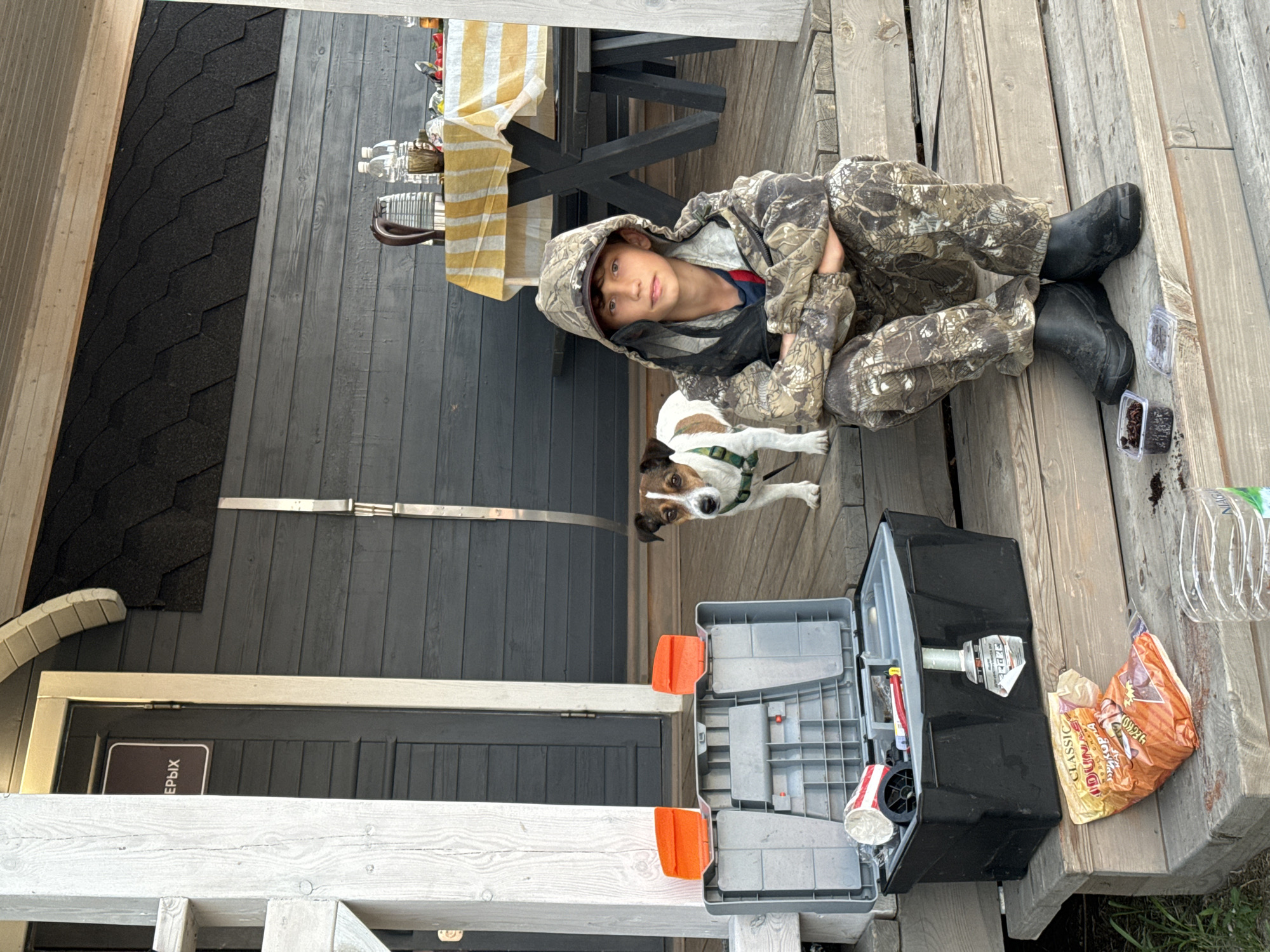 A young boy in camouflage clothing and black boots sits on a wooden deck next to a Jack Russell Terrier. The boy is wearing a hooded jacket, and his arms are crossed. The dog is looking towards the camera. A black open toolbox with orange latches is on the deck in front of them. To the right of the toolbox is a crushed orange bag of chips, and further to the right, a plastic water bottle. In the background, a dark gray wooden wall and a black shingled roof are visible. A striped yellow and white tablecloth is draped over a dark table. The scene appears to be outdoors, likely in a backyard or porch area. The lighting suggests it is daytime, possibly overcast. A sign on the door in the background has Cyrillic text that reads "ЕРЫХ". The overall mood seems relaxed and casual.