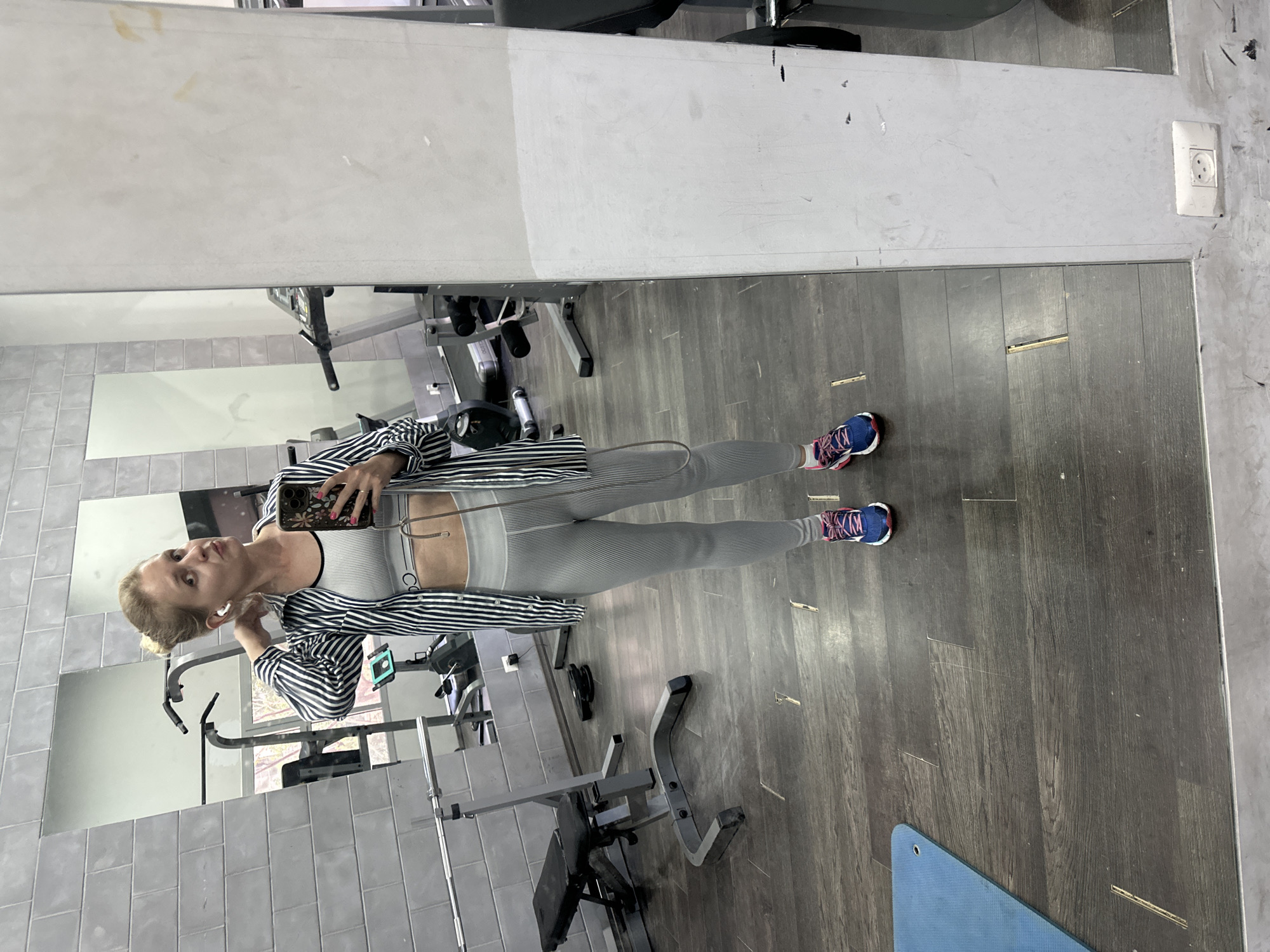 The image displays a young woman taking a mirror selfie within a gym or fitness center, located in Petah Tikva, Israel. She is reflected in a large mirror, standing centrally, and appears to be in her 20s or 30s. Her light-colored hair is styled back, and she is wearing a light grey or white sports bra with a visible "Calvin Klein" (or "CK") logo, light grey striped leggings, and a black and white striped long-sleeve shirt worn open. She has blue and pink/red athletic shoes on her feet and white wireless earbuds in her ears. In her left hand, she holds a smartphone with a floral-patterned case, directing her gaze towards it to capture the selfie.
The background, seen through the mirror's reflection, reveals a typical gym environment. Various pieces of grey and black exercise equipment, including weight benches and racks, are visible. The floor is dark wood laminate, exhibiting some signs of use. A blue exercise mat lies partially on the floor in the foreground. The walls feature light grey tiles and painted sections. A white European-style power outlet is present on the wall to the right. The mirrors themselves show some smudges and wear. The overall scene captures a moment of self-portraiture within a workout setting.