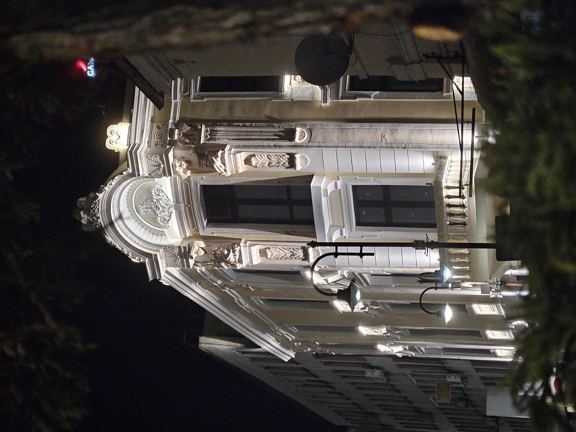 At night, the camera looks up at an ornate building facade in Burgas, Bulgaria. The building is lit with streetlights and has architectural details like columns, moldings, and decorative elements. A satellite dish is affixed to the exterior. The streetlights are on tall poles. A neon sign with the letters "GA" is visible in the top left corner. Branches of a tree frame the shot on the right side. The sky is dark.