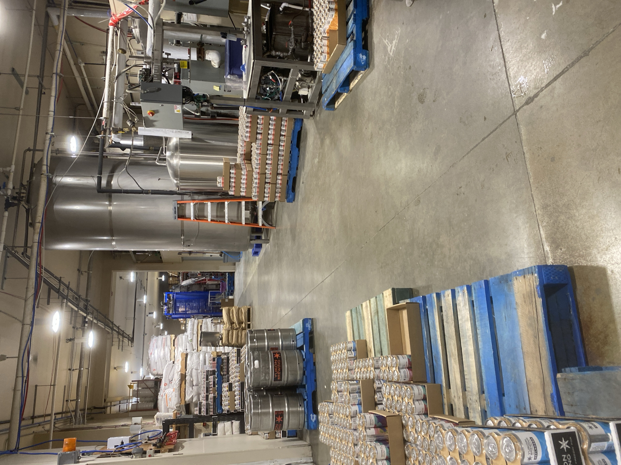 The image depicts the interior of a brewery or beverage production facility in Whitehorse, Canada. The environment is industrial, characterized by concrete flooring, exposed ceilings with piping and conduit, and large stainless steel tanks.
In the foreground, on the left side of the image, several pallets are stacked with cases of canned beverages. These cases are predominantly cardboard, and many of the cans are visible, displaying various colorful labels. Some of these cans are stacked in boxes, while others are arranged in rows on the pallets. One pallet in the immediate foreground has a portion of a blue wooden pallet visible.
Further into the scene, several large metal kegs, likely for beer or other beverages, are arranged on blue wooden pallets. These kegs appear to be made of stainless steel and bear some branding.
In the mid-ground, to the left, there are shelves stacked with bags of what appear to be raw ingredients, possibly grains or malt for brewing. There are also rows of smaller containers, potentially bottles or smaller cans, on shelves. A blue industrial machine is visible in the background behind these shelves.
Dominating the right side of the mid-ground and background are large stainless steel tanks, characteristic of brewing operations. Piping and various pieces of industrial equipment are connected to these tanks. An orange ladder is positioned near one of the tanks.
In the background, more industrial equipment and storage areas are visible, including more pallets of bagged materials and stacks of cardboard boxes. The overall lighting suggests an indoor industrial setting, likely during operational hours. There are no individuals clearly visible in the scene. The environment appears organized for production and storage.
