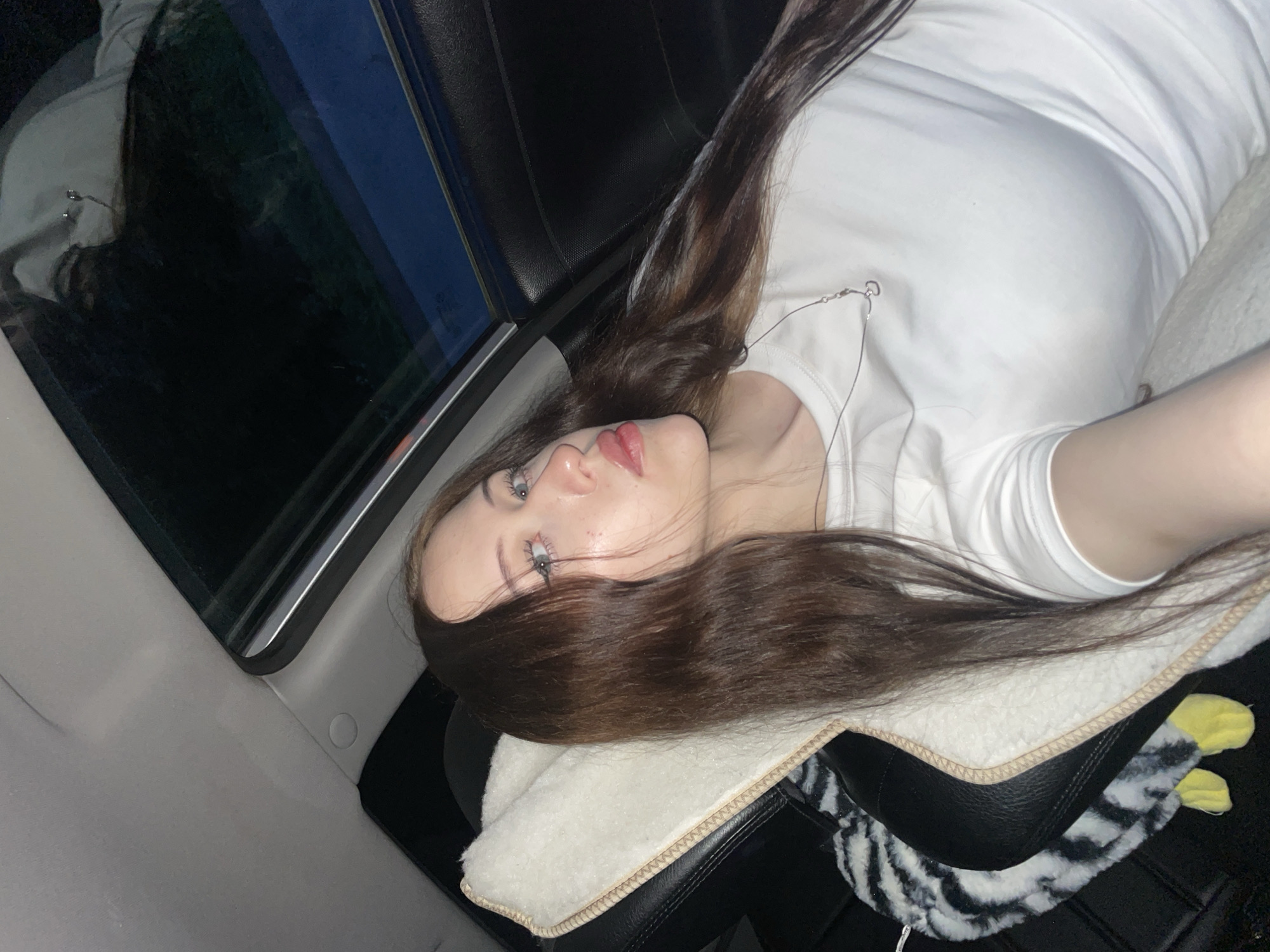 A young woman with long, dark brown hair and fair skin is reclining in the passenger seat of a car, looking towards the camera. She is wearing a plain white t-shirt and a silver necklace with a small heart pendant. Her eyes are light blue, and her lips are painted a light pink. She is resting her head on a cream-colored, fluffy headrest. The car's sunroof is open above her, revealing a dark, night sky. The interior of the car appears to be dark, with the black leather of the seat and a black and white striped pattern visible on another object in the foreground. The overall lighting suggests it is nighttime. The image does not contain any specific identifiable landmarks or features to pinpoint a location within the city of Gavrilovka, Russia.