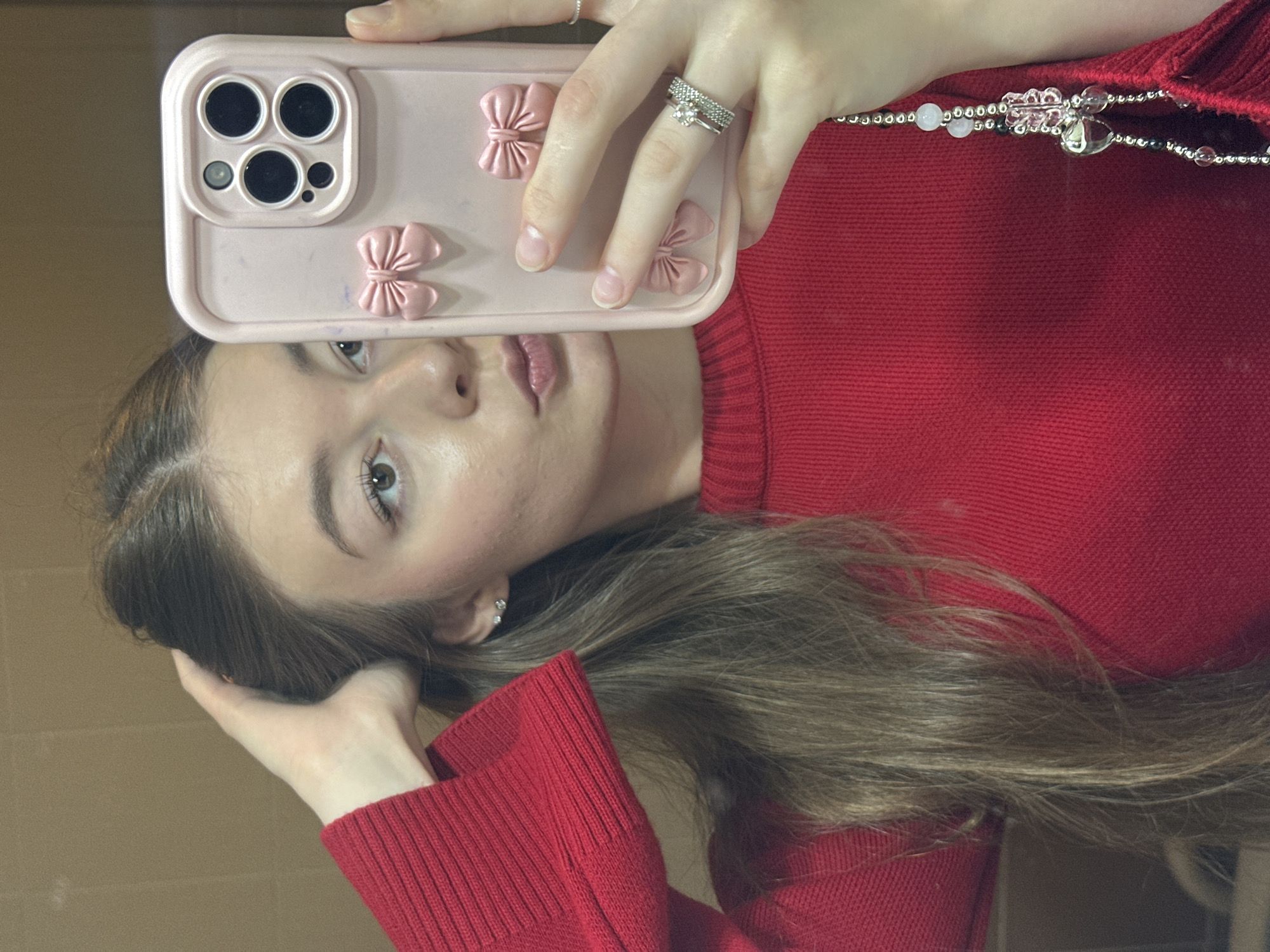 The original image displays a young woman taking a mirror selfie indoors, appearing rotated 90 degrees counter-clockwise. The woman, likely in Kazan, Russia based on provided context, has long, light brown hair, and wears a red ribbed knit sweater. She holds a light pink smartphone with a triple camera system, encased in a matching pink cover adorned with several small, three-dimensional pink bows.
Her right hand, holding the phone, displays two rings on her ring finger: one a simple band, the other set with a clear stone. Her left hand rests casually near her hair. A multi-beaded necklace, featuring silver, light pink, clear, and dark beads, along with a pink flower-shaped charm and a silver heart charm, is visible draped over her right shoulder. A small, clear stud earring is visible in her left ear.
The woman's face is reflected in the mirror; she has light-colored eyes and her lips appear to have a glossy finish. Her expression is neutral. The background behind her reflection is a plain, light brown or beige wall, suggesting an indoor setting. No specific time of day or weather conditions can be discerned, and there is no visible text in the image.