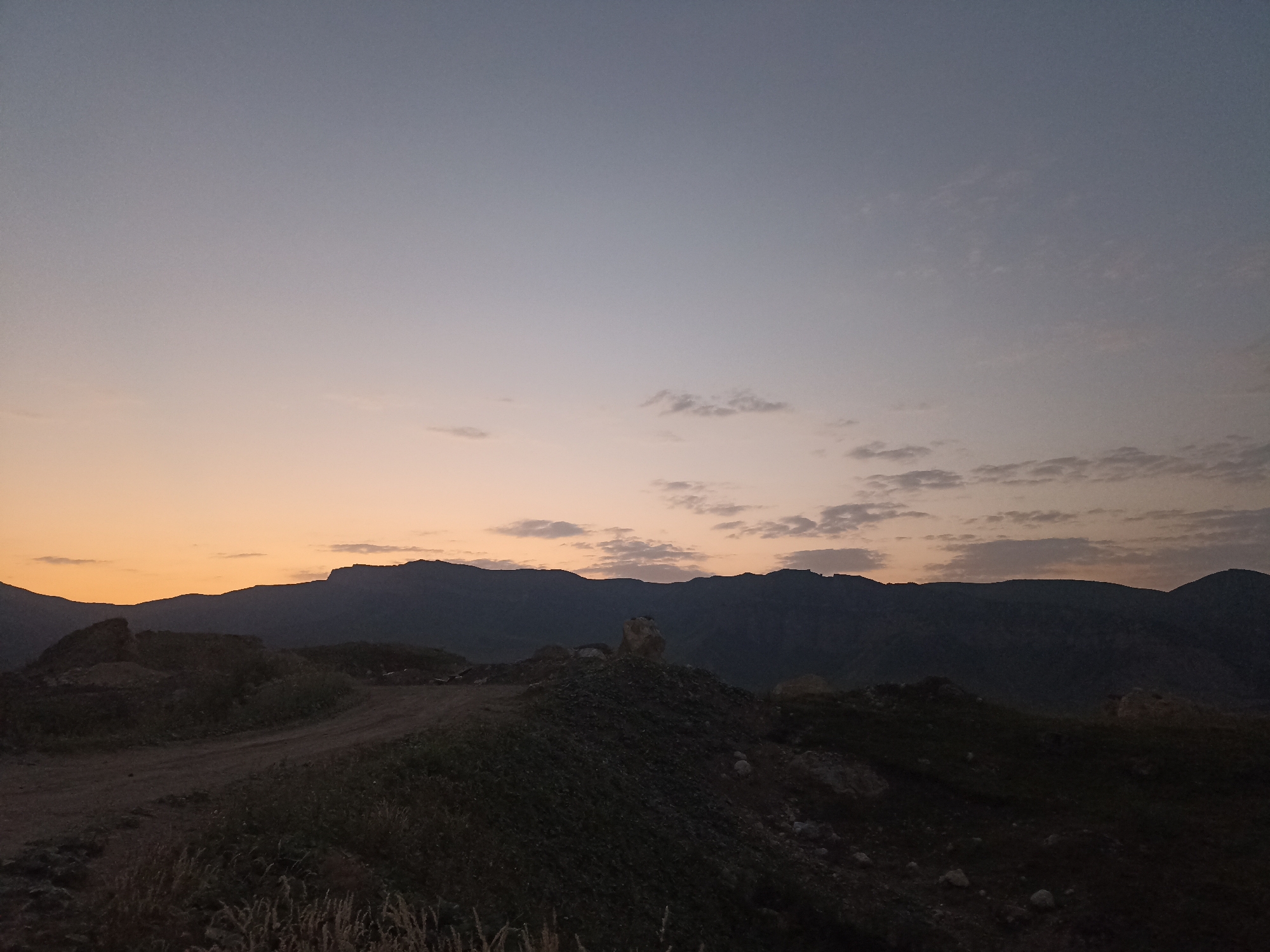 The image depicts a serene, twilight landscape with rolling hills and a dusky sky. In the foreground, a dirt path winds through a hilly terrain covered in sparse vegetation and rocks. The hills in the midground are dark and silhouetted against the horizon, with a jagged mountain range forming the backdrop. The sky transitions from a soft orange hue near the horizon to a pale blue-gray overhead, with scattered wispy clouds. The scene is devoid of people or any man-made structures, suggesting a remote, natural setting. The overall atmosphere is calm and tranquil, characteristic of dusk in a rural or mountainous area. The location is specified as Kuzun, Azerbaijan.