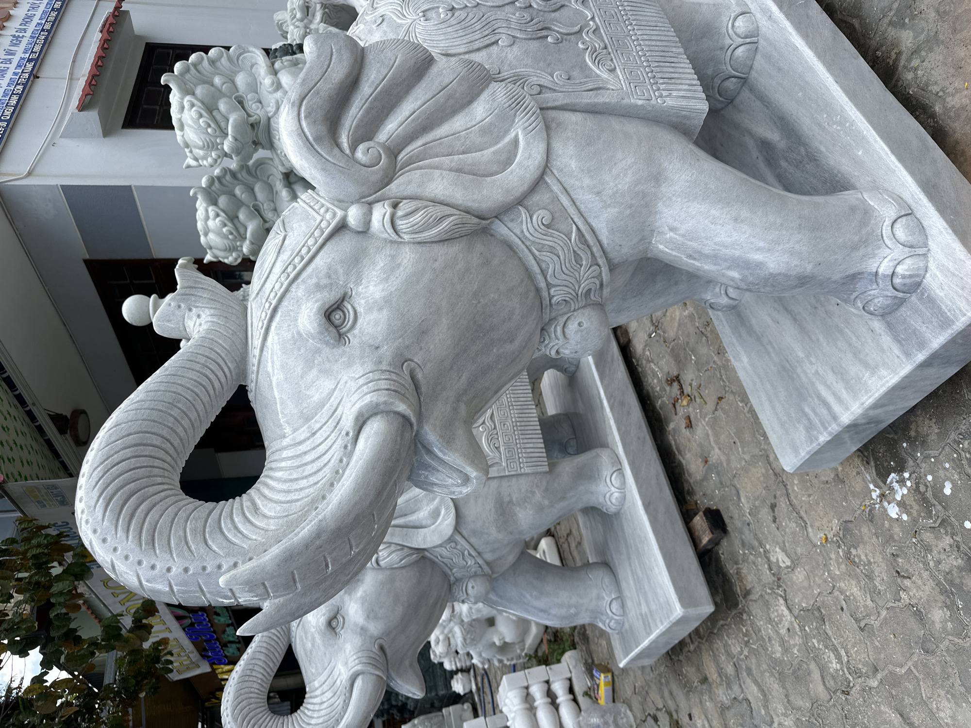 This image depicts a street-level view outside a stone carving shop in Da Nang, Vietnam. The shop appears to be located on a paved street with visible signs in Vietnamese. The foreground is dominated by two intricately carved marble elephant statues. The elephant on the right is in sharper focus, showing detailed carvings on its tusks, trunk, ears, saddle, and legs. The elephant on the left is partially visible behind it. In the background, more stone carvings, possibly other animal statues and architectural elements, are stacked and displayed. A building with windows and a tiled roof is visible behind the statues. The overall scene suggests a commercial establishment specializing in stone sculptures, situated in an urban Vietnamese environment.