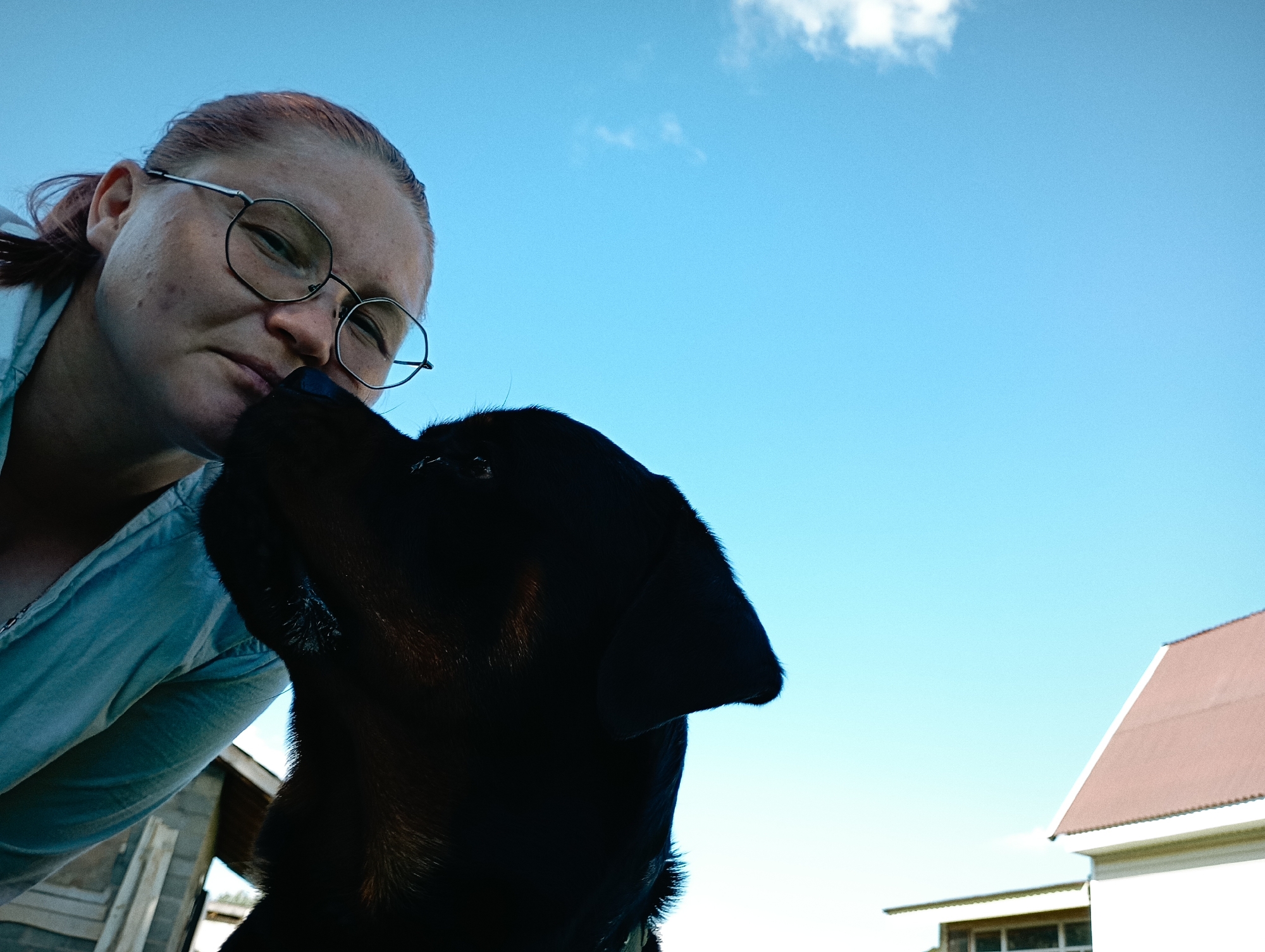 A low-angle outdoor photograph shows a person wearing glasses and a light blue collared shirt, looking down at a black dog. The person's hair is tied back, and they appear to be in their late teens or early twenties. The dog's head is in the foreground, obscuring part of the person's face. The background consists of a clear blue sky with a few white clouds, and a portion of a building with a reddish-brown roof. The image captures a moment of interaction between the person and the dog, possibly in a rural setting. There is no identifiable information that places the scene specifically in the city of Ryabki, Russia.