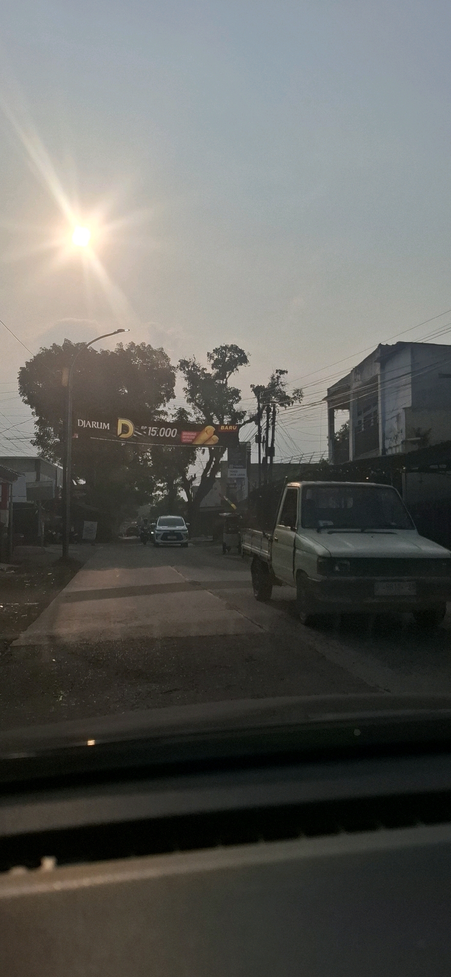 In Sukasari, Indonesia, a street scene is captured at sunrise or sunset. The sun, a bright lens flare, is positioned in the upper left quadrant of the sky, casting a warm, golden light. A large banner advertising "DIARUM D RP 15.000 BARU" hangs from a lamppost, partially obscured by trees. A white pickup truck is parked on the right side of the road, facing forward. A white sedan is visible on the left side of the road, also facing forward. The road surface is paved, with shadows cast across it from the surrounding trees and buildings. In the background, there are various buildings and utility poles with a dense network of wires. The overall atmosphere is tranquil, with the warm light of the sun creating a serene mood.