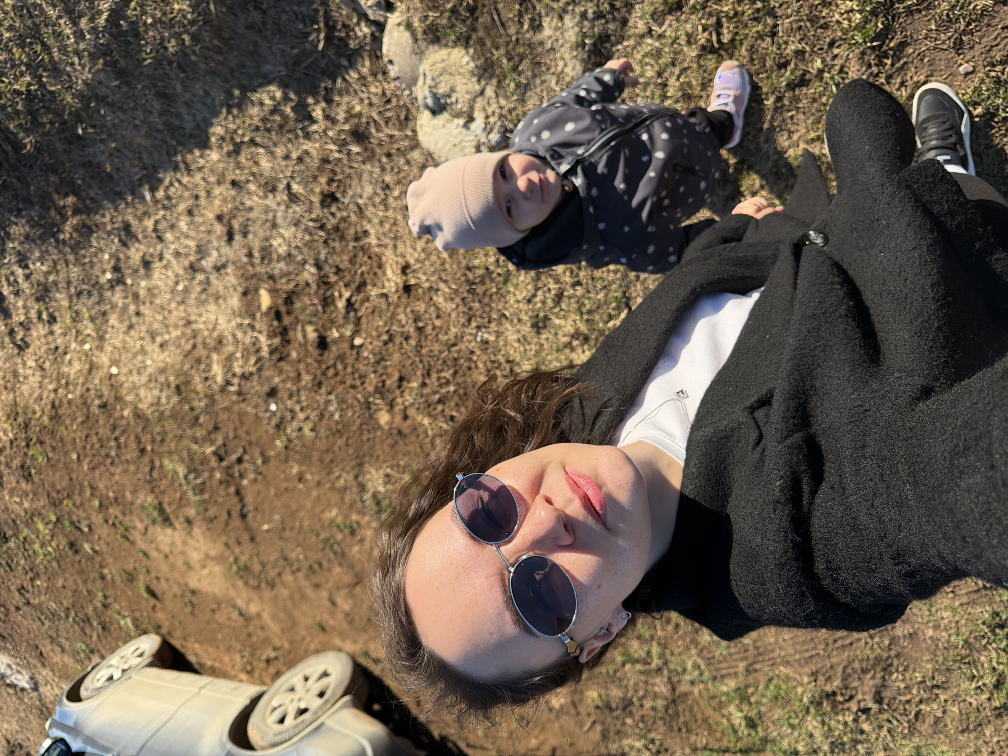 The image depicts a woman wearing sunglasses and a black coat lying on the ground with a child in a dotted coat and beanie. They are outdoors, possibly in a rural or park-like setting with dry, grassy ground. A white car's wheels are visible in the lower left corner. The location is Большой Букор, Russia.