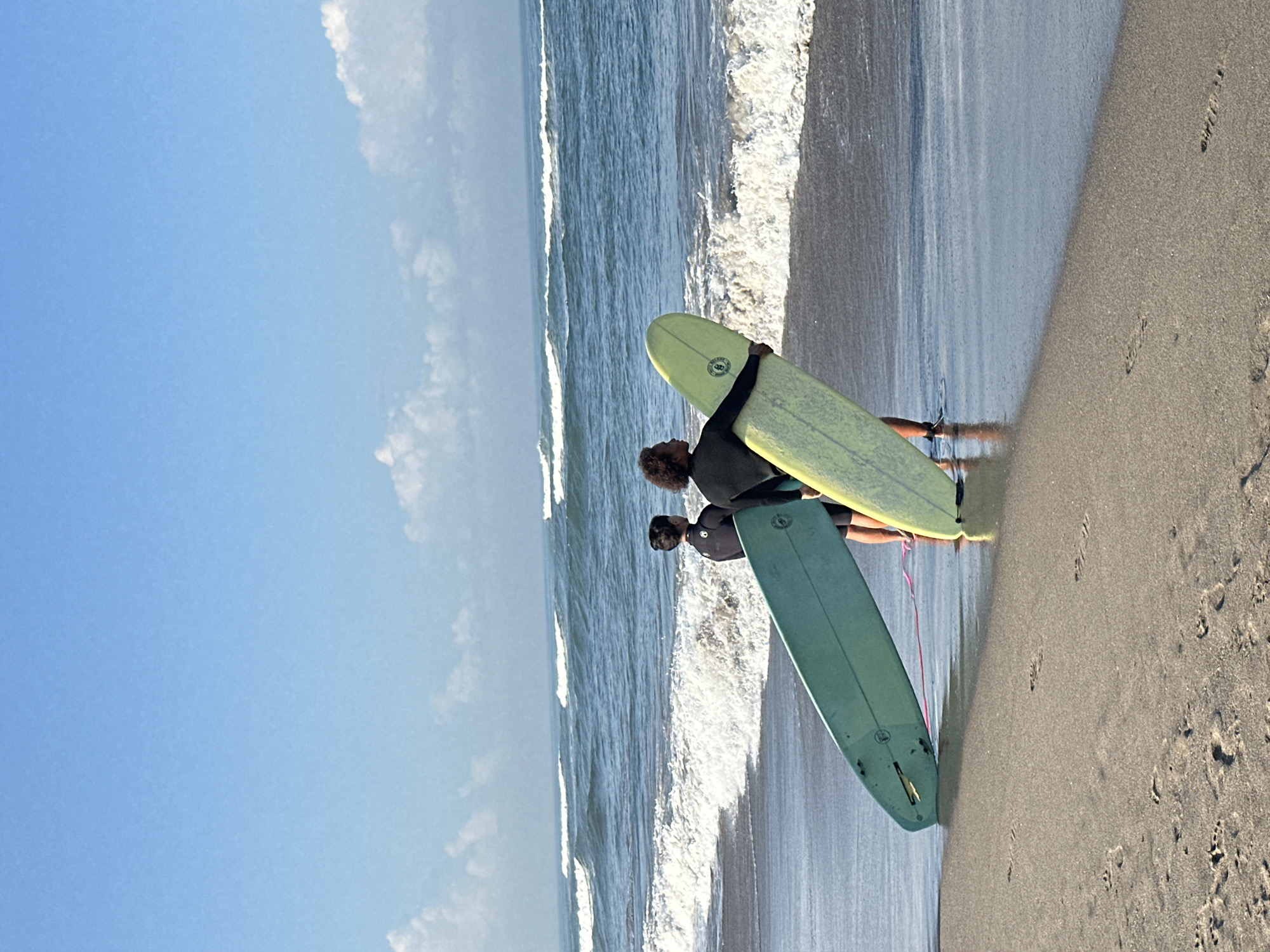 The image shows two individuals standing on a beach in North Kuta, Indonesia, holding surfboards. The person on the left is wearing a black wetsuit and holding a light teal surfboard. The person on the right is wearing a black wetsuit and holding a light yellow surfboard. They are both facing the ocean, which has moderate waves breaking on the shore. The sky is clear and blue with scattered white clouds. The sand on the beach is wet and appears to have footprints. The overall scene depicts a beach environment suitable for surfing.