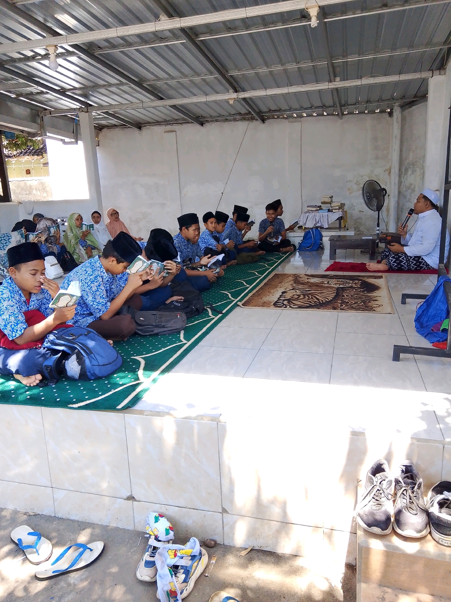 In an open-air covered structure in Kuripan Utara, Indonesia, a group of young boys and a teacher are engaged in an educational activity. The boys, dressed in blue patterned shirts and black peci caps, are seated on a green mat on the floor, each holding and reading a book. Some are looking intently at their books, while others are looking towards the teacher. The teacher, wearing a white robe and cap, stands on a raised platform on the right side of the frame, holding a microphone and addressing the boys. A fan is positioned behind him. Various bags are placed on the floor near the boys. In the foreground, outside the structure, several pairs of footwear are scattered, including flip-flops and sneakers. The environment appears to be a functional, possibly outdoor classroom setting, with a tin roof overhead supported by pillars.