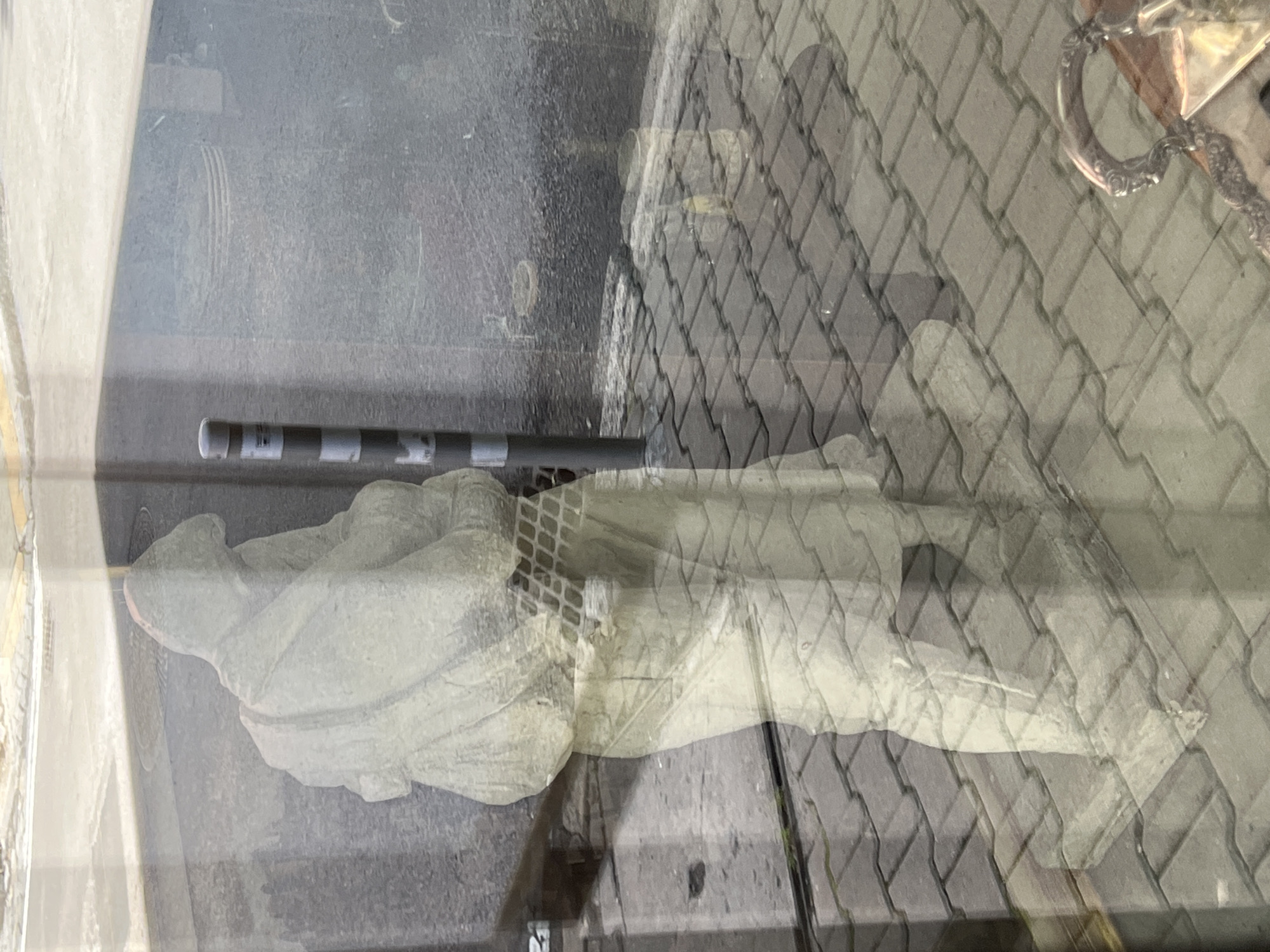 The image shows a reflection of a statue or sculpture of a person on a paved surface, likely viewed through glass. The statue appears to be light-colored and depicts a figure with a hooded garment, with a bag or backpack slung over one shoulder. The figure is in a standing position, with one leg slightly forward. The background reflection shows a street scene with a bollard featuring black and white stripes, a manhole cover, and a textured asphalt or concrete surface. The ground in the foreground of the reflection is paved with interlocking gray bricks. A portion of what appears to be an ornate metal object, possibly a tray or handle, is visible in the lower right corner of the image. The overall lighting suggests it is daytime. The prompt indicates the location is Sofia, Bulgaria, but there are no explicit visual cues in the image to confirm this.