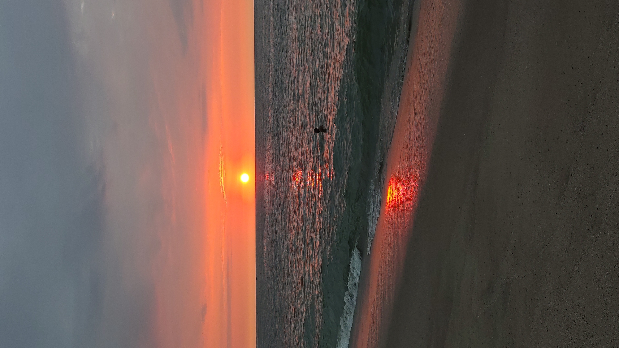 A beach scene at dusk features a body of water reflecting the setting sun, with a person wading in the water. The sky is a gradient of orange, red, and gray, suggesting a sunset. The beach is sandy and dark, with the water's edge reflecting the sun. The person in the water is a dark silhouette, making their identity unclear. The location is likely Bocapán, Peru, implied by the prompt's request.