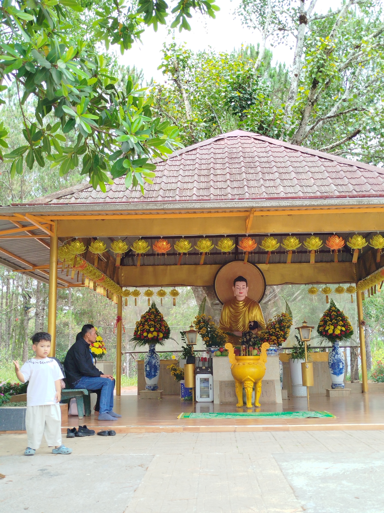 The image depicts a scene at a Buddhist shrine or temple in Da Lat, Vietnam. A young boy, dressed in white clothing and blue sandals, stands in the foreground with his left hand raised. Behind him, an adult male sits on a green bench. The shrine features a golden Buddha statue at the rear, adorned with flower arrangements and lights. In front of the statue, there is a yellow incense burner. The structure has a brown tiled roof and is decorated with lotus-shaped lanterns. The surrounding environment includes trees and greenery, suggesting the temple is located in a natural setting.