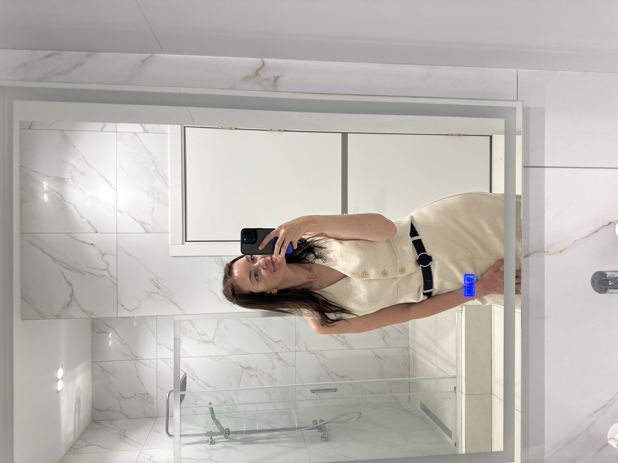 A woman is taking a selfie in a bathroom mirror. The bathroom features white marble-patterned walls and a glass-enclosed shower. Above the shower, a white cabinet is visible. The woman is wearing a light-colored dress with a black belt. The reflection shows a section of the bathroom including a sink with a faucet. No information is available on whether this location is Прикамский, Russia.