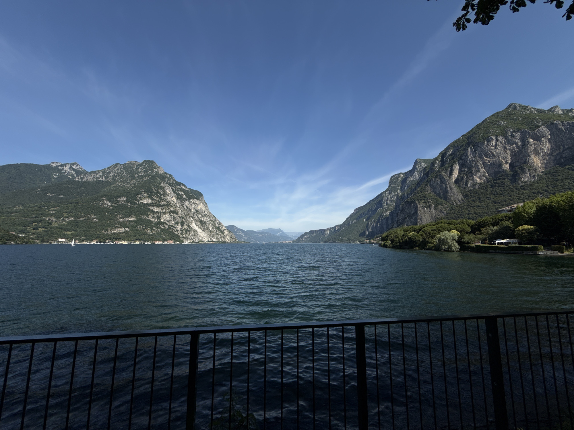 A wide-angle, eye-level outdoor shot shows a serene lake flanked by steep, tree-covered mountains under a clear blue sky with thin white clouds. The dark blue water of the lake is slightly rippled. In the foreground, a black metal railing runs horizontally across the bottom of the frame. On the far side of the lake, a thin strip of land is visible, dotted with buildings and lush green trees. In the distance, more mountains are visible, creating a layered effect. A single sailboat is present on the lake. The overall scene suggests a peaceful, natural landscape.