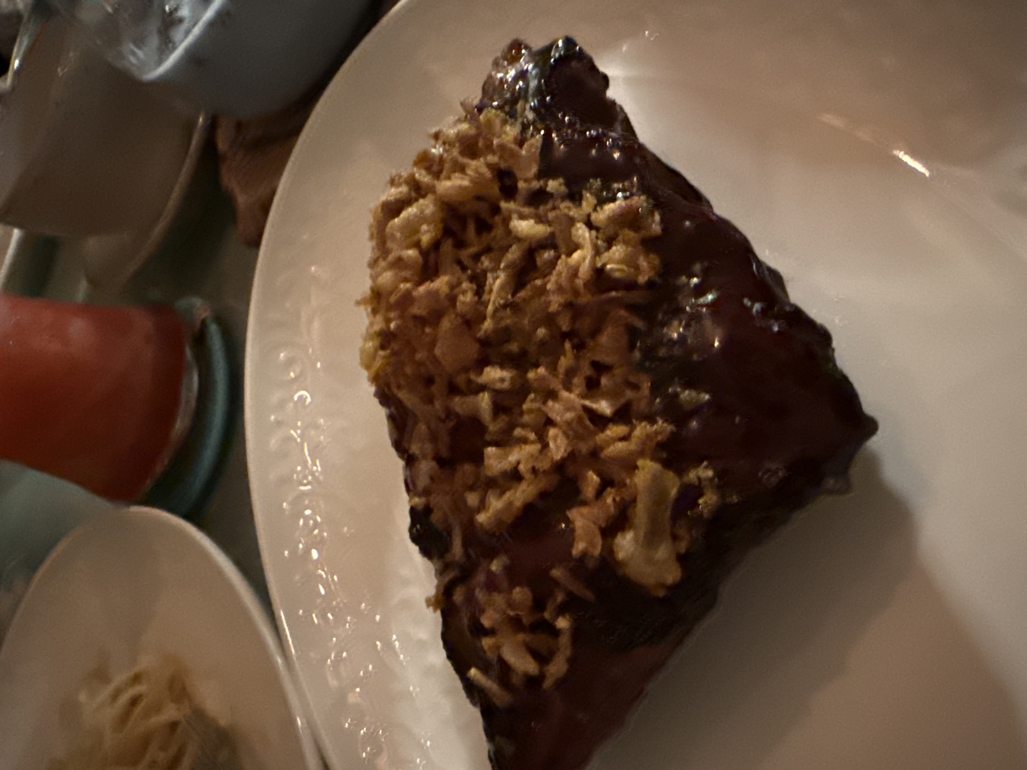 A piece of meat, likely ribs, coated in a dark sauce and topped with crispy fried onions is presented on a white plate. The meat is glistening, and the onions are scattered across its surface. The plate is part of a larger table setting, with other plates and drinks visible in the background. The lighting suggests an indoor dining environment. There is no information about the location in Feodosia, Ukraine.