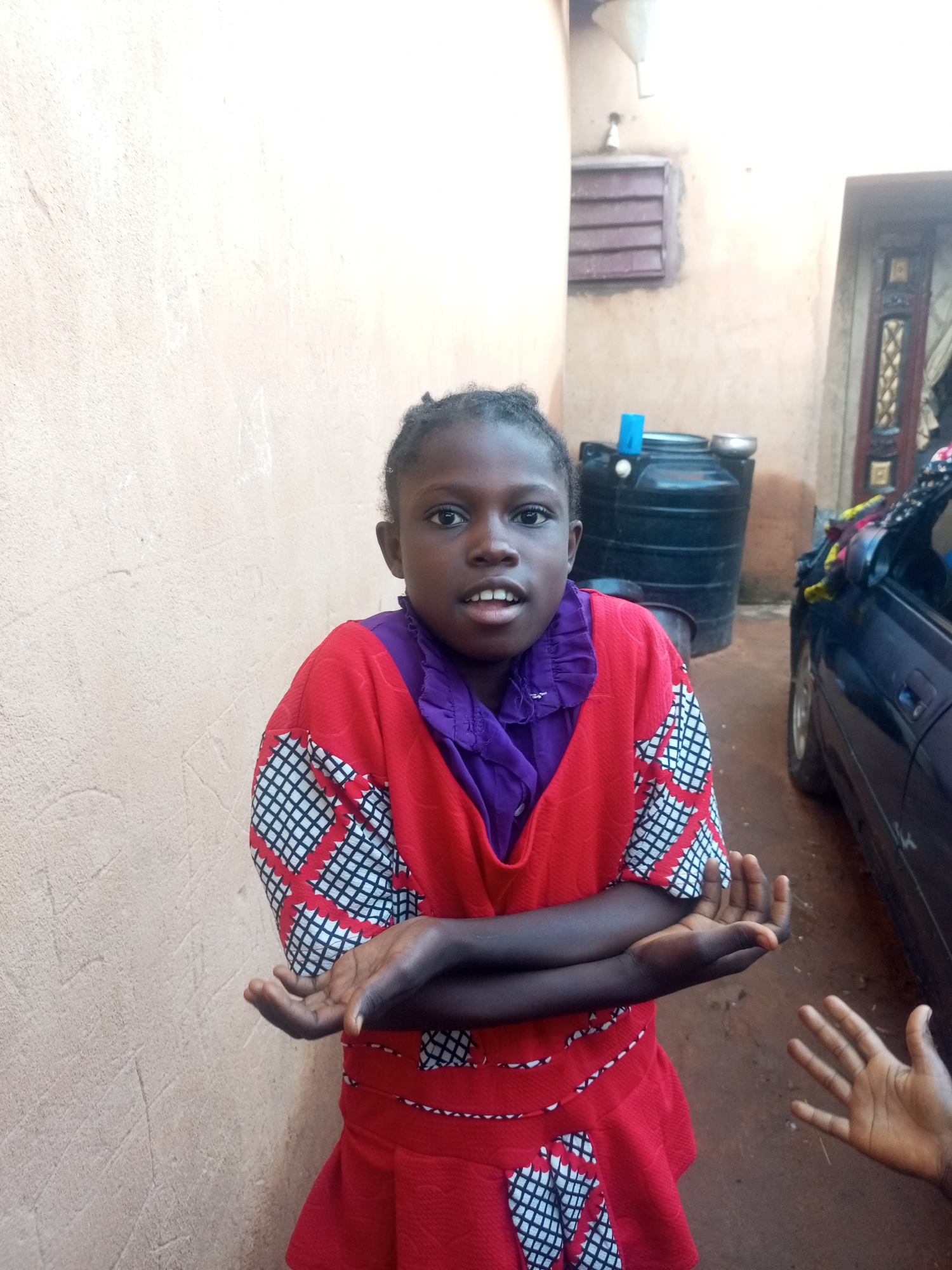 A young girl stands in front of a beige wall in Tempure, Nigeria. She is wearing a red and purple dress with a patterned design. Her arms are crossed in front of her chest, and her hands are open, palms facing upward. Her mouth is slightly open as if she is speaking. Behind her, a black water tank and a portion of a blue car are visible. The environment appears to be an outdoor, residential area with a rough, dirt ground.
