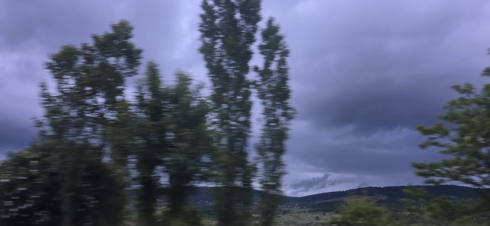 The image depicts a landscape viewed through a moving window, characterized by significant motion blur. Tall, slender trees with dark green foliage occupy the foreground, appearing as vertical streaks due to the speed of movement. The sky is heavily overcast with dark, ominous purple-gray clouds, suggesting stormy weather or a twilight setting.
In the mid-ground, a range of hills or mountains is visible, partially obscured by the heavy clouds and atmospheric haze. These landforms are covered in dark vegetation, and faint indications of scattered structures, possibly houses, can be seen along their lower slopes. The overall lighting is dim and diffused, contributing to a dramatic and somewhat somber mood. There are no identifiable people, animals, or visible text in the image.