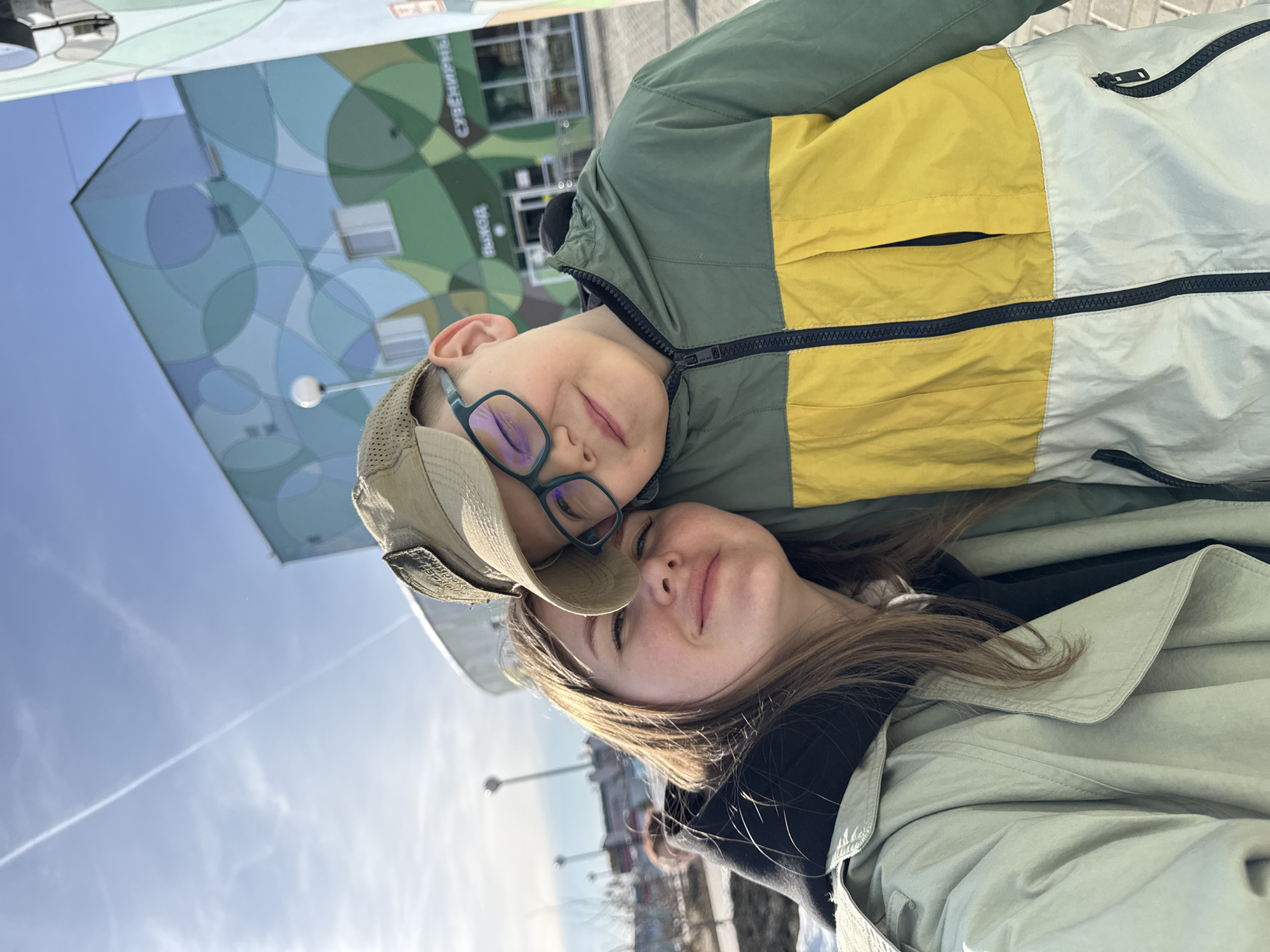 Two people, a boy and a girl, are posing for a selfie. The boy wears a cap and glasses, while the girl has long brown hair. They are dressed in jackets. In the background, a building with a patterned facade is visible, along with streetlights and a blue sky. The building has the Russian word "Северный" (Severny) on it. Based on this, the location is likely Perm, Russia, as "Северный" translates to "Northern" and there is a neighborhood by that name in Perm.