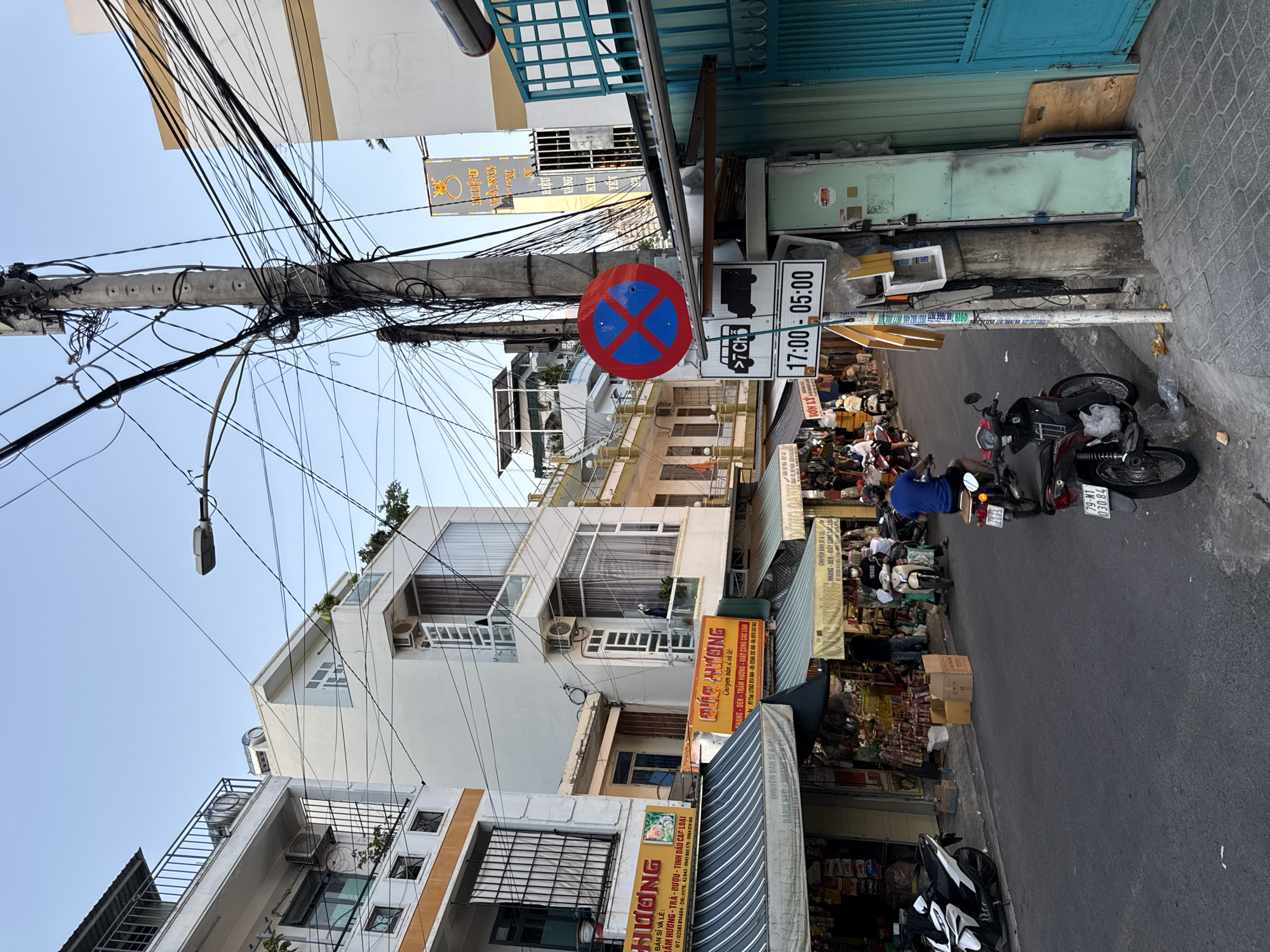 The image depicts a street scene in Nha Trang, Vietnam, characterized by narrow buildings, a bustling street, and a tangle of electrical wires overhead. On the left, a multi-story building with balconies and air conditioning units is visible. Adjacent to it, another building features shops with striped awnings selling various goods. A street sign indicates that parking is prohibited from 5:00 PM to 5:00 AM. Several motorbikes are parked on the side of the road, and people are seen walking and riding them. The street is paved, and the buildings are closely packed, typical of an urban environment. A tall utility pole, laden with electrical wires, stands prominently in the center of the image, a common sight in Vietnamese cities. The sky is clear and appears to be late afternoon or early evening.
