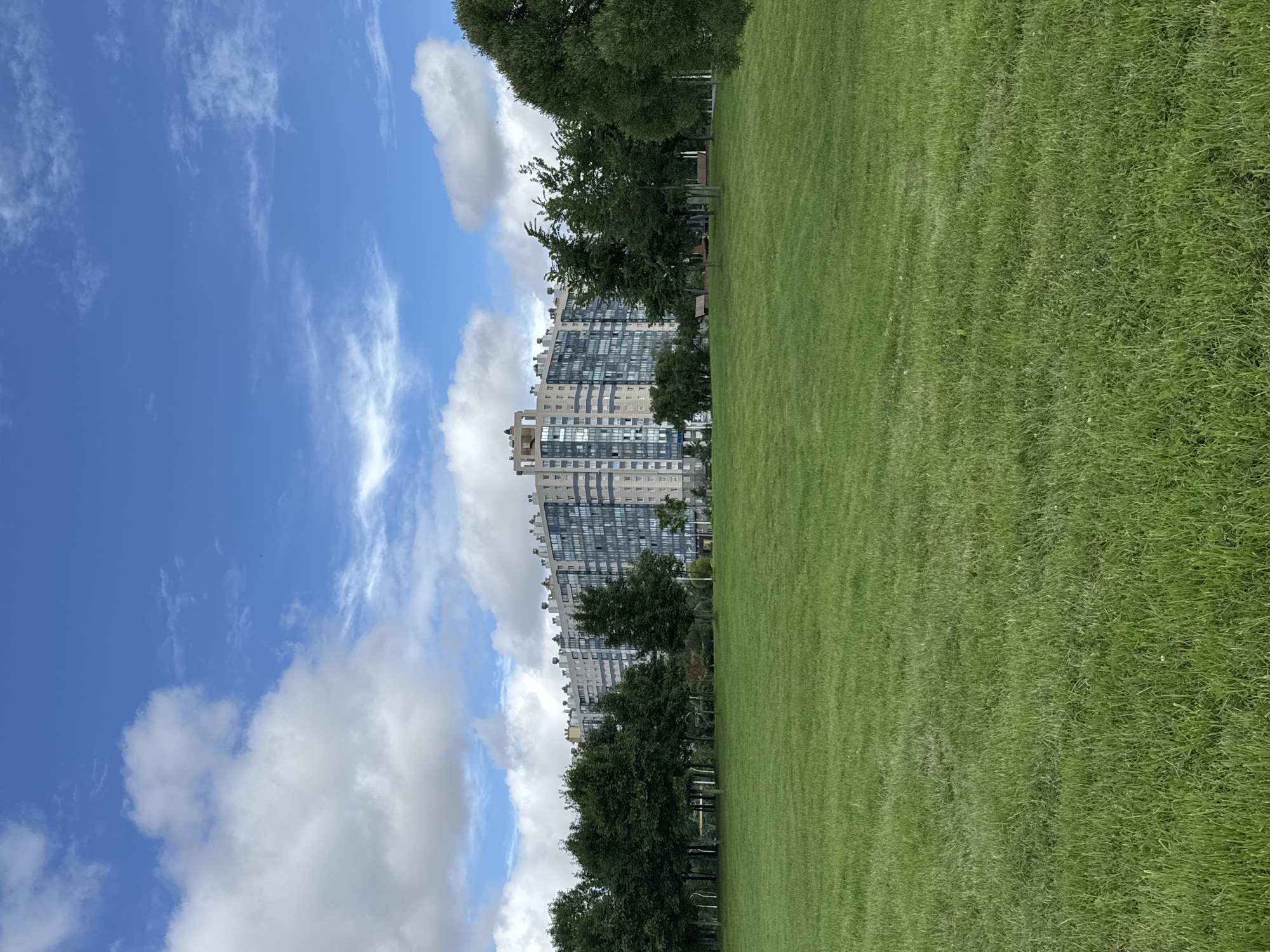 The image depicts a vast, vibrant green lawn in the foreground, stretching towards a line of lush green trees. Behind the trees, a large, modern apartment complex with a light-colored facade and numerous windows rises against a bright blue sky dotted with white, fluffy clouds. The apartment building has a distinctive tiered design at the top and appears to be a residential area. The scene is set in the city of Sinyavino, Russia, likely a park or open recreational space adjacent to residential buildings.