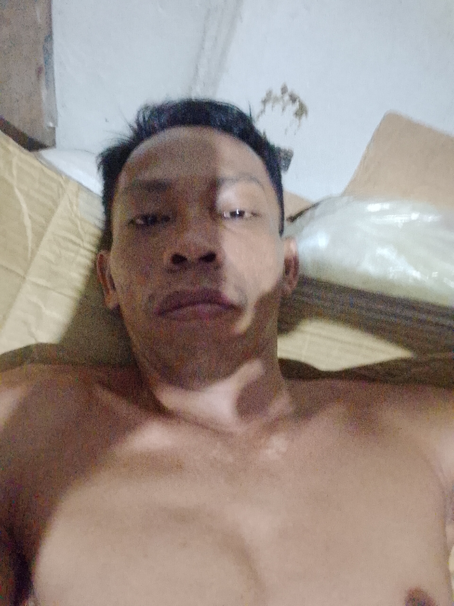 A shirtless man is lying down indoors. He is positioned at a slight upward angle, looking directly into the camera. His head is resting on what appears to be a cushion or pillow. The background is partially obscured, but includes what may be cardboard boxes and a white wall. There is evidence of stains or discoloration on the wall. The location is Bukit Ketok, Indonesia.