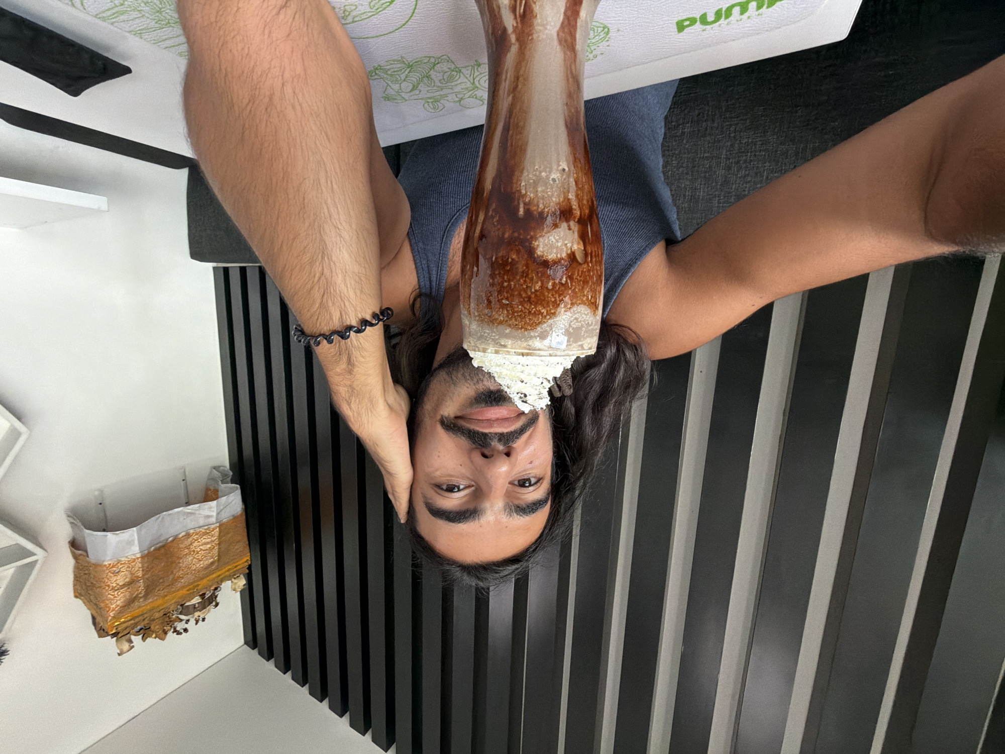 A man with long, dark hair and a mustache poses for a selfie in front of a dark, slatted wall. He is wearing a blue tank top and has his left hand on his head. In front of him is a tall glass filled with a chocolate milkshake topped with whipped cream. A black, spiral bracelet is visible on his right wrist. On the wall behind him, a small, golden altar with offerings is mounted. The scene appears to be indoors, possibly in a cafe or restaurant in Mengwi, Indonesia, given the altar decoration.