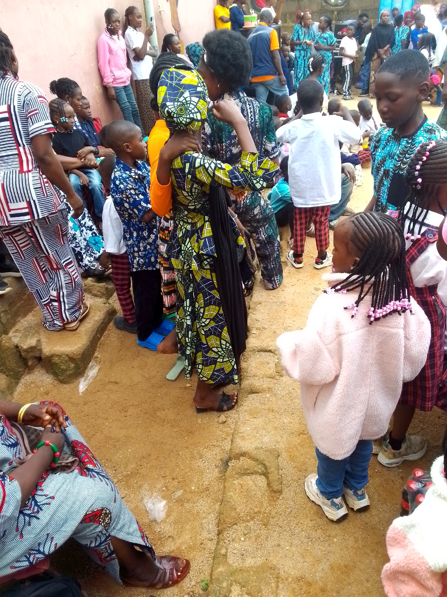 In an outdoor, dirt-and-sand environment, a group of people, predominantly children and young women, are gathered. Many are wearing brightly colored clothing, including patterned dresses and shirts. Some individuals have elaborate hairstyles. In the foreground, a young girl with braided hair adorned with pink beads wears a fluffy pink jacket and blue jeans, standing in white sneakers. To her left, another child, possibly a boy, is seen from the back wearing a blue and white patterned shirt and red checkered pants. A woman in a blue, white, and red patterned dress is visible on the left side of the frame. Further back, a larger group of people stand together, some wearing matching blue and white patterned outfits. The background suggests a somewhat informal gathering, possibly a celebration or community event, with indistinct structures and other individuals. The overall impression is one of a lively, communal gathering in a warm climate.