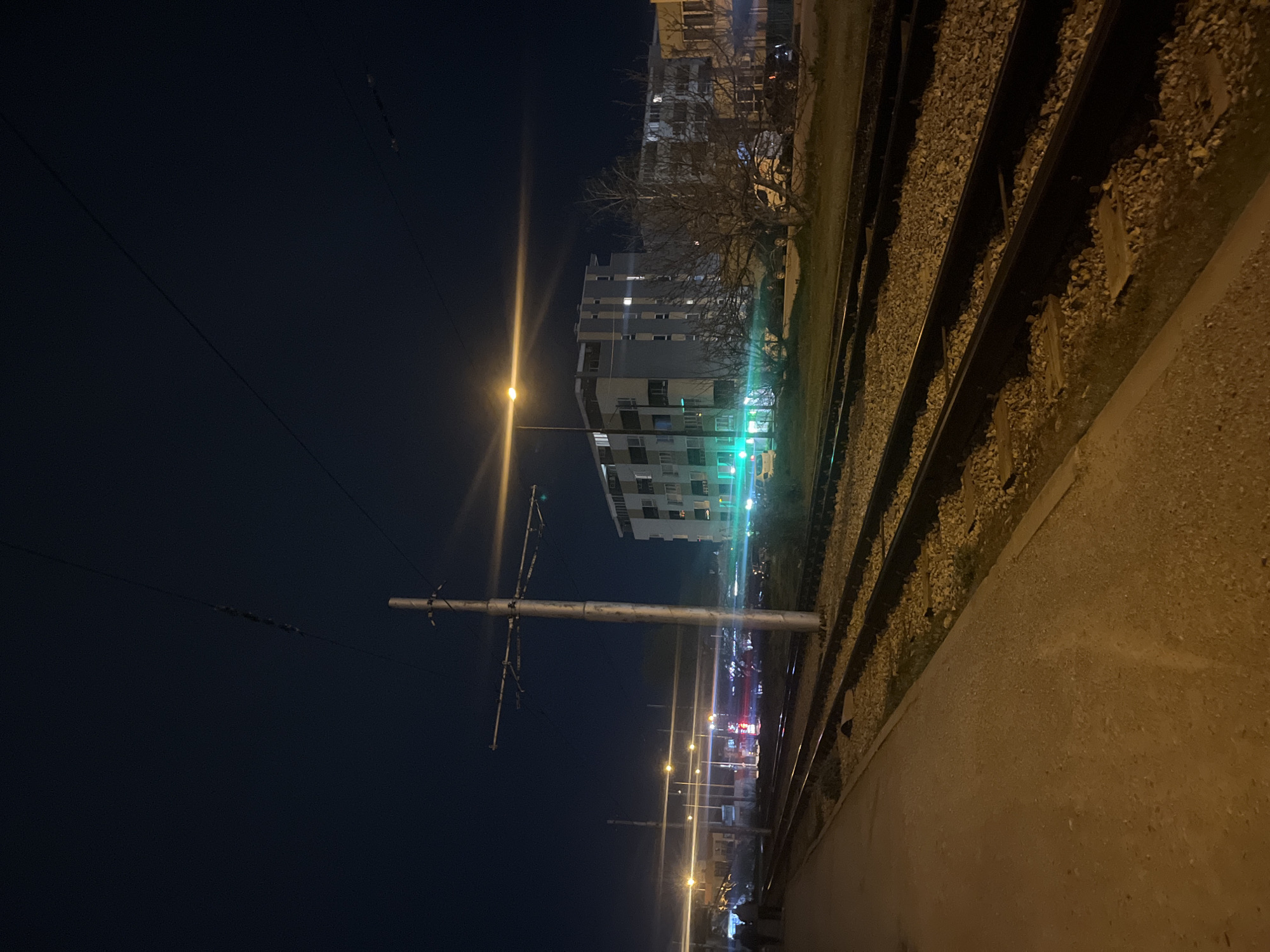 The image shows a night scene in Sofia, Bulgaria, along a section of railway tracks. The perspective is from the side of the tracks, looking towards buildings lit by streetlights and apartment lights. There is a utility pole with wires extending into the dark sky. The tracks themselves are bordered by gravel and a concrete or asphalt edge. The overall atmosphere is dimly lit with contrasting dark and light areas.