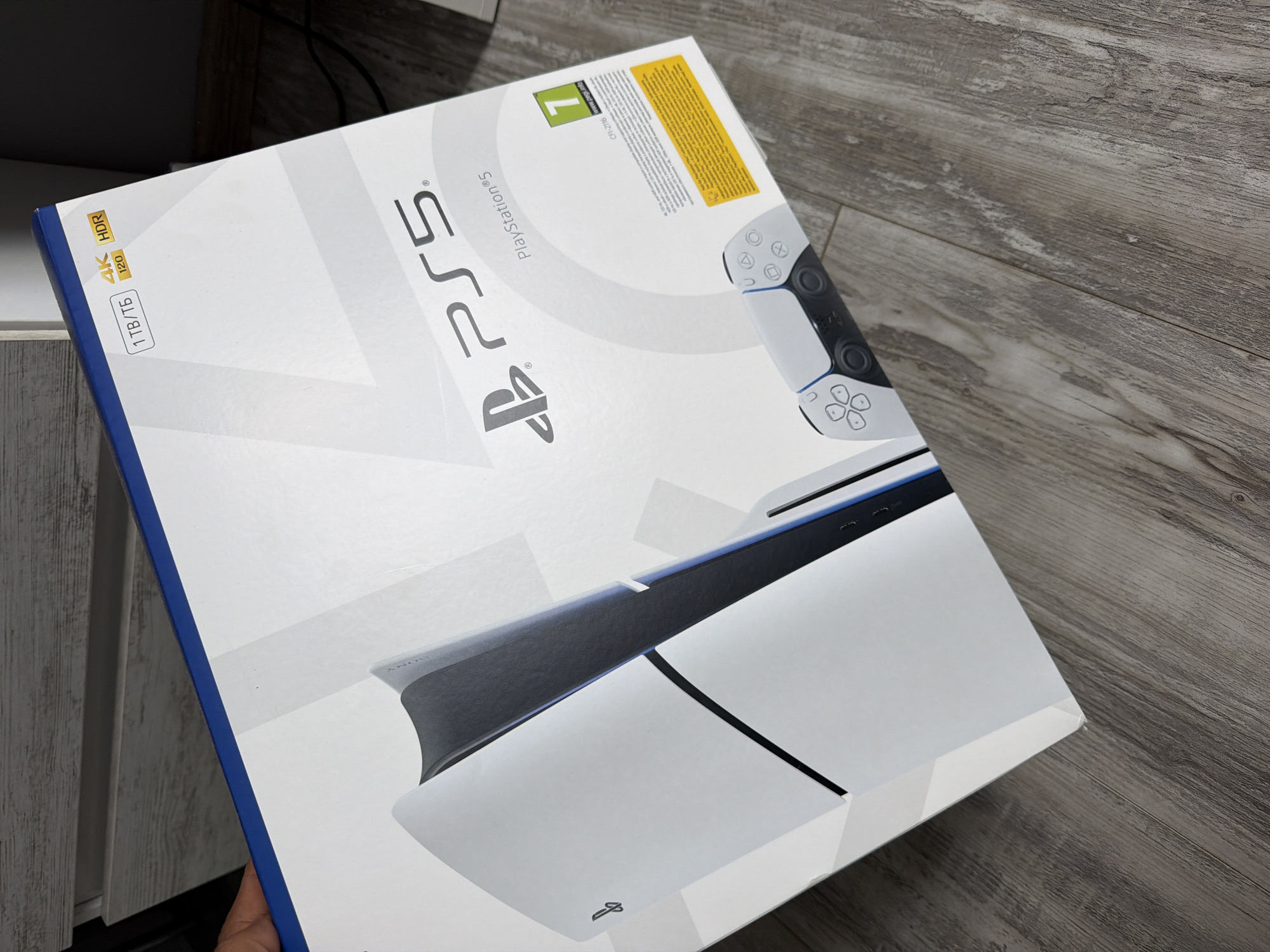 This media file features a PlayStation 5 console box held by a person over a light-colored wooden or laminate floor. The box is predominantly white with a blue edge visible on the left side, showcasing a large image of the PlayStation 5 console (disc version) and a DualSense controller.
Notable details on the box include:
- The prominent "PS5" and "PlayStation®5" branding.
- Specifications like "1TB/ТБ" (indicating 1 terabyte of storage), "4K 120", and "HDR" (High Dynamic Range), suggesting its visual and performance capabilities.
- A green PEGI age rating icon with the number "7" and "www.pegi.info", indicating its suitability for ages seven and up.
- A model number "CFI-2116".
- A yellow rectangular block containing small, illegible text, likely legal or product information in various languages.
- The faint "SONY" branding is visible on the console depicted on the box.
- A small PlayStation logo symbol is also present on the front of the console shown in the illustration.
A portion of a hand is visible at the bottom left, holding the box, implying a person is either presenting it or has just acquired it. The background consists of a textured, light-greyish wooden floor and a dark, possibly wooden, furniture piece or shelf to the left. The scene is indoors, likely a home or retail environment in Dabene, Bulgaria, given the provided context. The lighting suggests an indoor setting without specific time-of-day indicators.