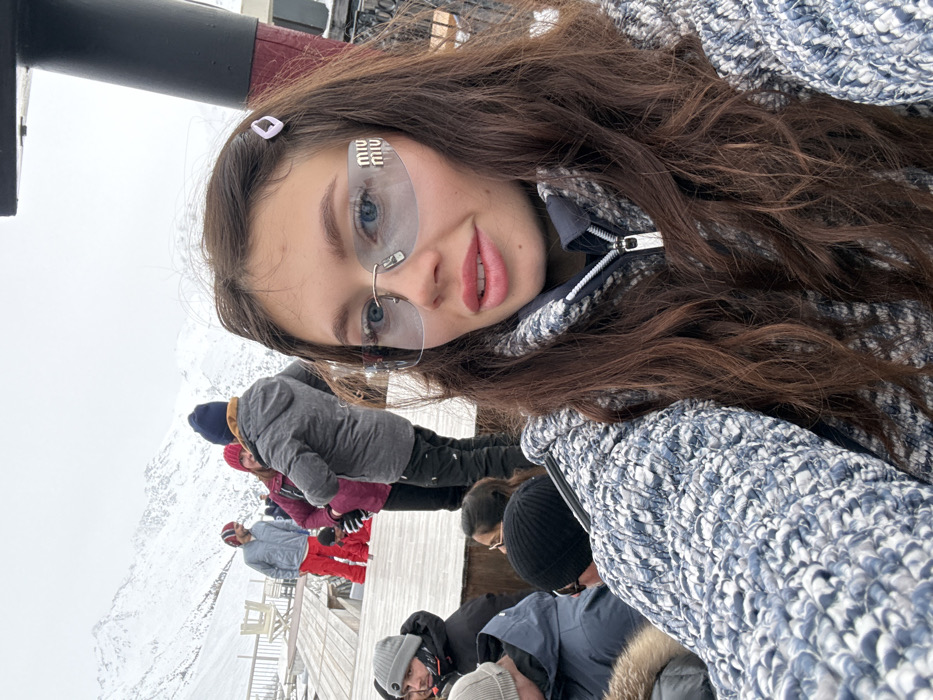 A young woman poses in a selfie, showcasing her long brown hair, light blue eyes behind designer sunglasses, and a gray and blue knit jacket. Behind her, a group of people stand on a wooden platform in a snowy, mountainous landscape. The background is slightly blurred, emphasizing the woman as the focal point of the image.