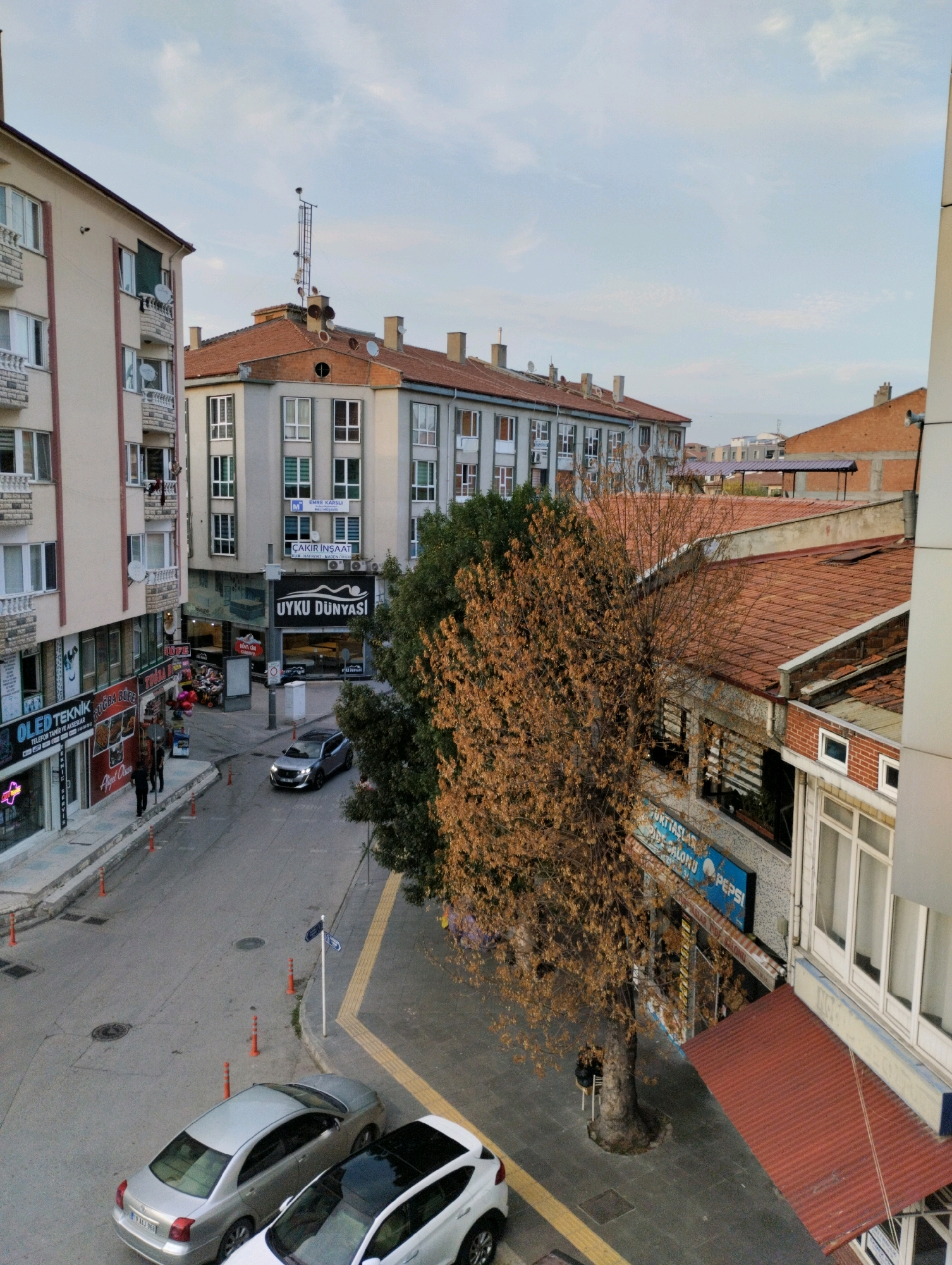 The image captures an urban street scene in Çorum, Turkey. In the foreground, two cars are parked on the right side of the road: a silver sedan and a white car with a black roof. A street runs through the middle of the frame, with pedestrian sidewalks lining both sides.
On the left side of the street, a multi-story building features shops on its ground floor with signs that read "OLED TEKNİK," "Telefon Tamir Ve Aksesuar," and "RUFE YORGAN." Further down the street, another building has a prominent sign for "UYKU DÜNYASI" (Sleep World). The buildings appear to be of concrete construction with multiple windows.
On the right side of the street, a large tree with golden-brown autumn leaves stands prominently. Behind the tree, a multi-story building with red-tiled roofs is visible, housing various businesses, including one with a blue sign that appears to say "PEPSİ."
The sky above is partially cloudy, suggesting it's daytime. The street is paved, and traffic cones are placed along the sidewalks. The overall impression is a typical urban commercial area.