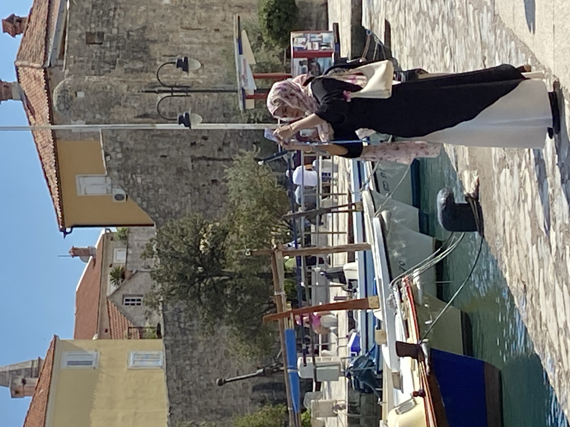 The image depicts a woman wearing a headscarf and carrying a white bag, standing at the edge of a pier, possibly using a mobile phone or taking a photo. The pier is adjacent to a harbor filled with boats, with visible mooring posts. The background includes the old town of Budva, Montenegro, recognizable by its aged stone buildings with terracotta-tiled roofs and historic architecture. A streetlight is visible against the backdrop of the stone buildings. The weather appears to be sunny, as indicated by the clear sky.
