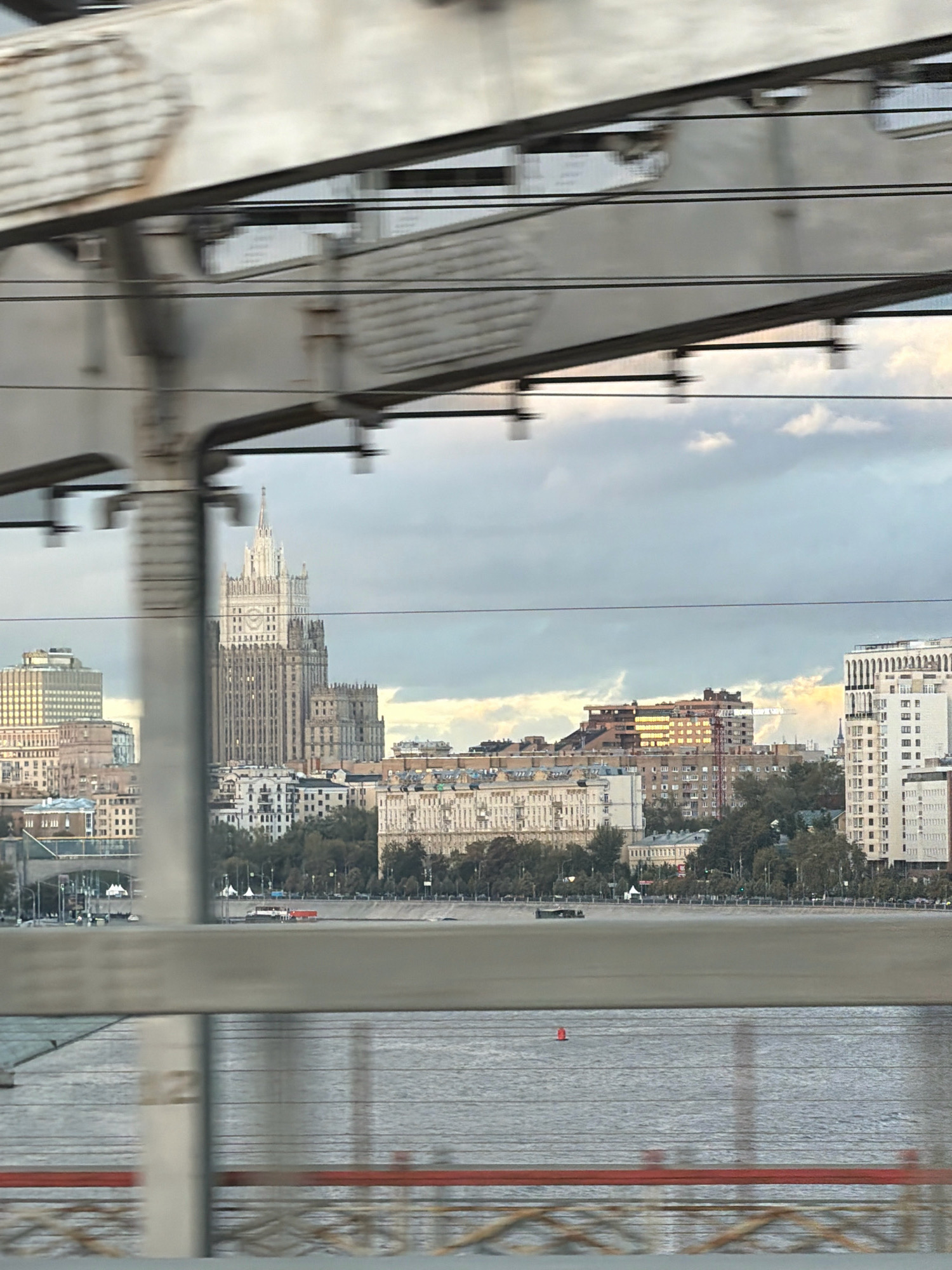 This image, captured from a moving vehicle, shows a panoramic view of the Moscow skyline, dominated by the Stalinist Empire-style architecture characteristic of the city. In the center, the imposing Ministry of Foreign Affairs building stands tall, its distinctive spire reaching towards a cloudy sky. Surrounding it are various other buildings of different heights and architectural styles, some appearing older and more ornate, others modern.
The foreground is partially obscured by the blurry, metallic structure of a bridge or overpass, with visible girders and cables creating a layered effect. Below the skyline, a wide river, likely the Moskva River, stretches across the scene. The water's surface is calm, with a few small boats or barges visible. The overall impression is of a bustling urban environment, viewed from a slightly elevated perspective, with the motion blur adding a sense of dynamism. The lighting suggests it is late afternoon or early evening, with the sun casting a warm glow on some of the buildings.