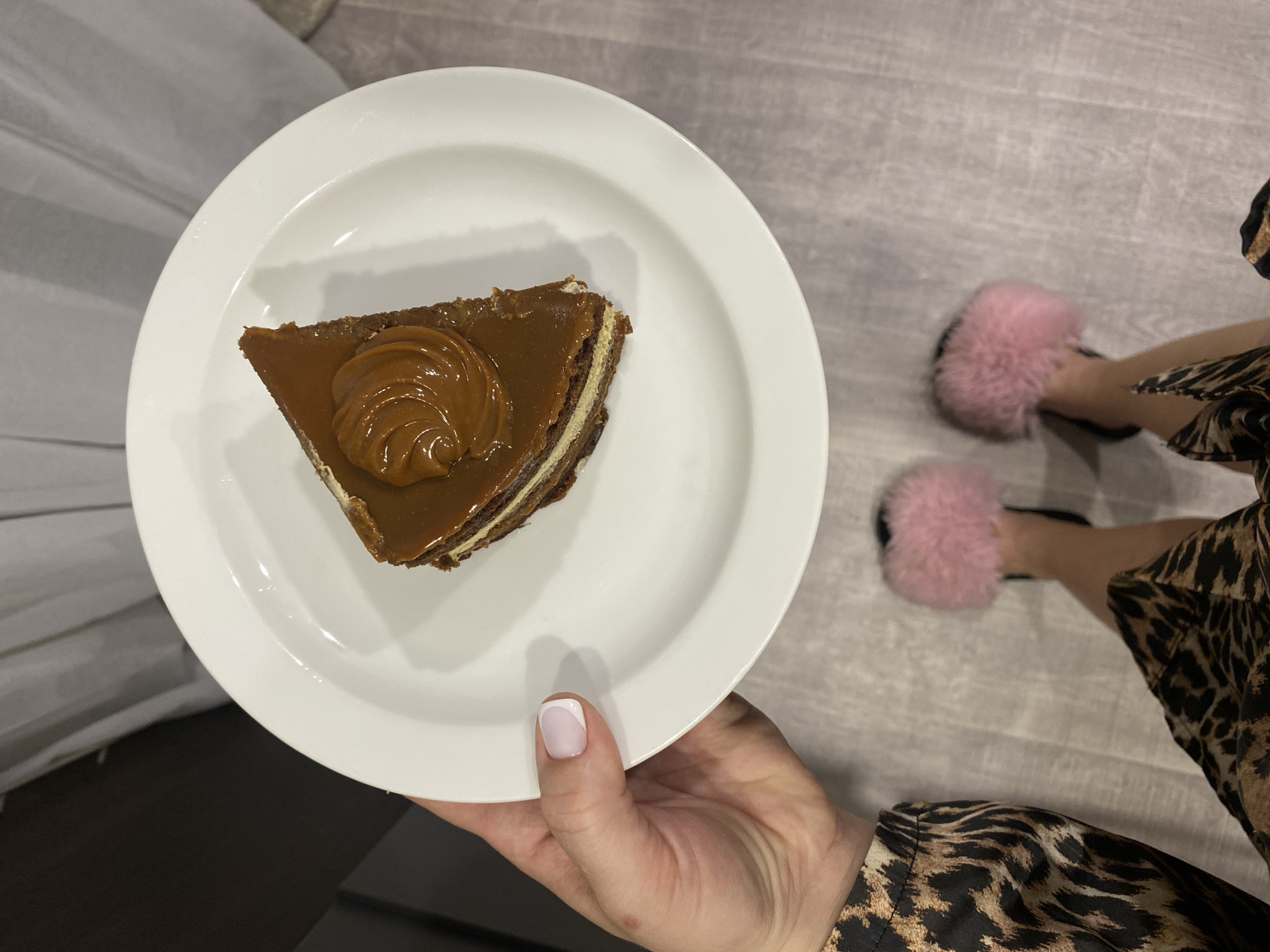 A top-down view shows a person holding a white plate with a slice of cake. The cake is triangular with multiple layers and a brown caramel topping decorated with a swirl of caramel. The person has a manicured hand with a white-tipped nail on their thumb. The person is wearing a leopard print robe or dress and pink fuzzy slippers. The setting appears to be indoors. The location is not discernible from the image.