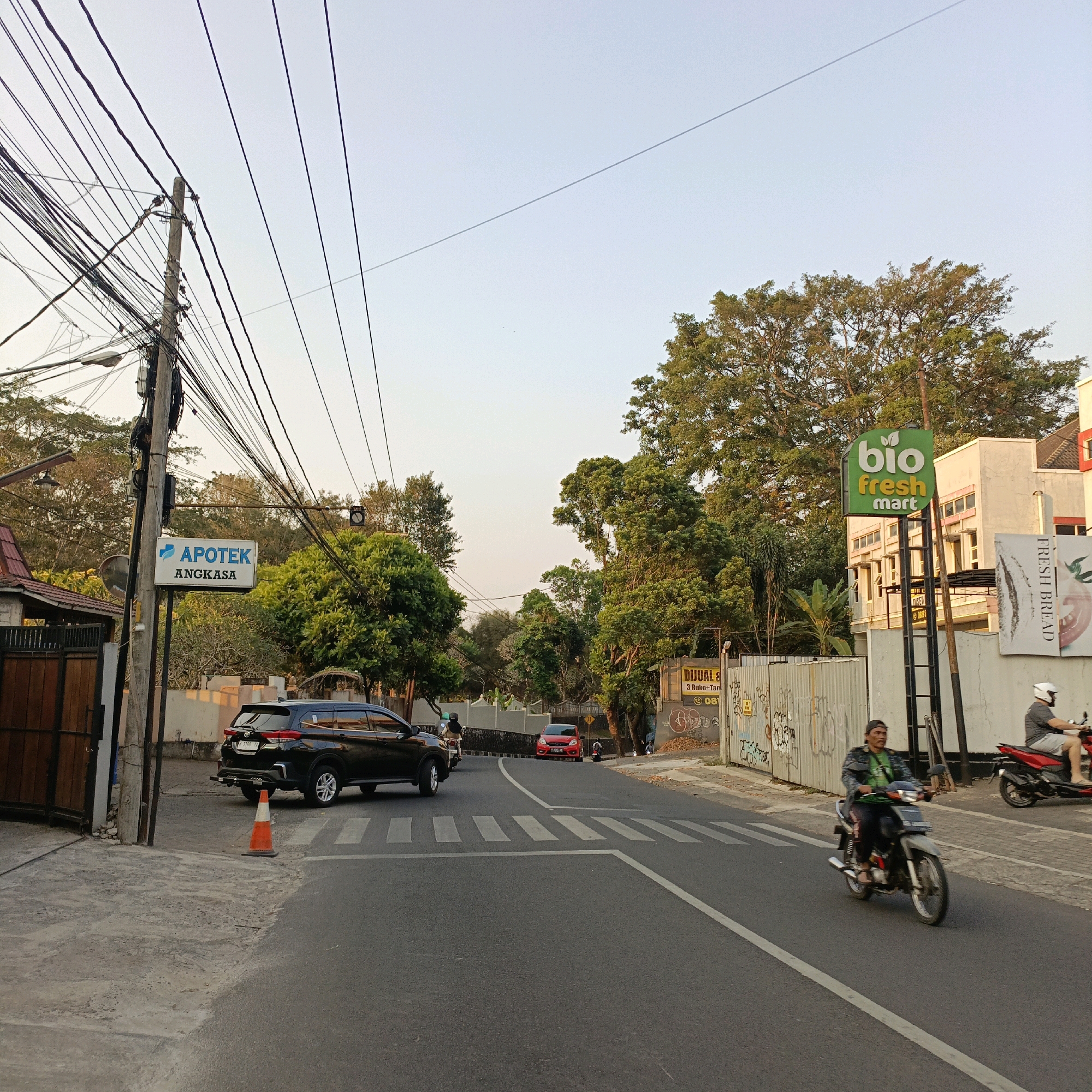 The image captures a street scene in Salatiga, Indonesia, under a clear sky with soft, late afternoon light. The foreground shows an asphalt road with a pedestrian crossing marked by white stripes. A black SUV is parked on the left side of the road, near a bright orange traffic cone. To the left, a wooden gate and a building with a red roof are visible.
A prominent pole on the left holds a sign for "APOTEK ANGKASA" (Angkasa Pharmacy), with numerous electrical wires entangled around it, creating a dense network overhead. Further down the road, on the right side, a green sign for "bio fresh mart" is mounted on a pole. Adjacent to this, a building with multiple windows stands. A large, leafy tree dominates the mid-ground on the right.
Several individuals are depicted in motion. A man on a motorcycle is riding towards the viewer on the right side of the road. Another person on a red motorcycle is visible in the distance to the right. A red car is stopped at the pedestrian crossing in the middle of the road. The environment is characterized by a mix of urban infrastructure, residential elements, and lush vegetation. The overall atmosphere is that of a typical day in a bustling Indonesian town.