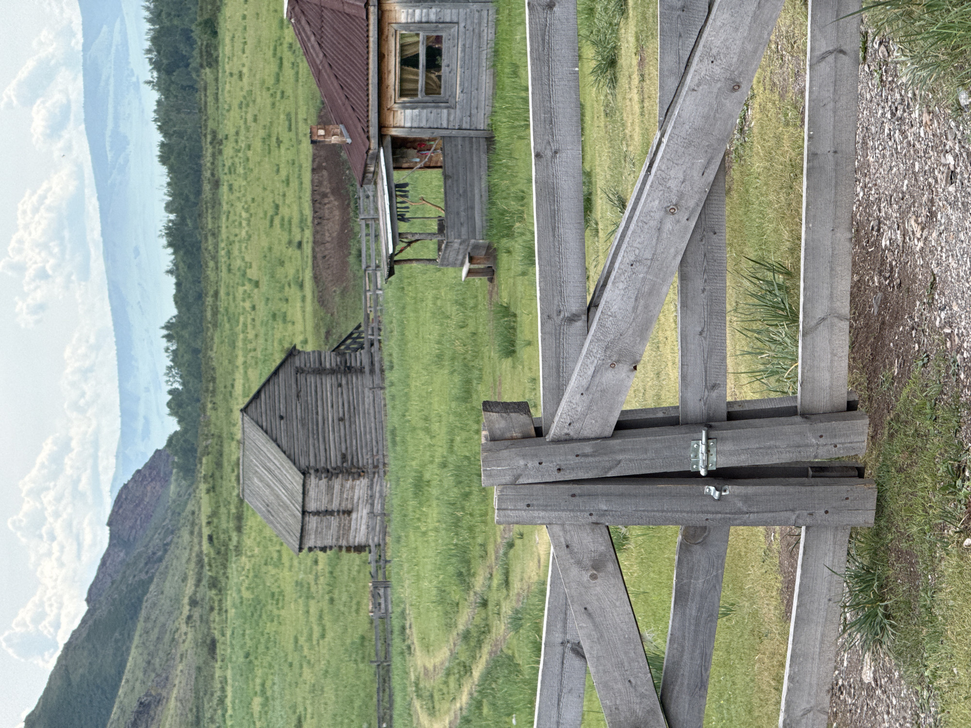 The image shows a wooden fence in the foreground, with a gate secured by a metal latch. Beyond the fence, a grassy field stretches out, with a dirt path winding through it. In the mid-ground, there are two rustic wooden structures. The one on the left is a log cabin, and the one on the right appears to be a small house with a porch and a red corrugated metal roof. In the background, a wooded area can be seen, followed by rolling hills and mountains under a cloudy sky. The scene is set in a rural, natural environment, possibly the village of Anchyl-Chon, Russia, characterized by its remote and pastoral landscape.
