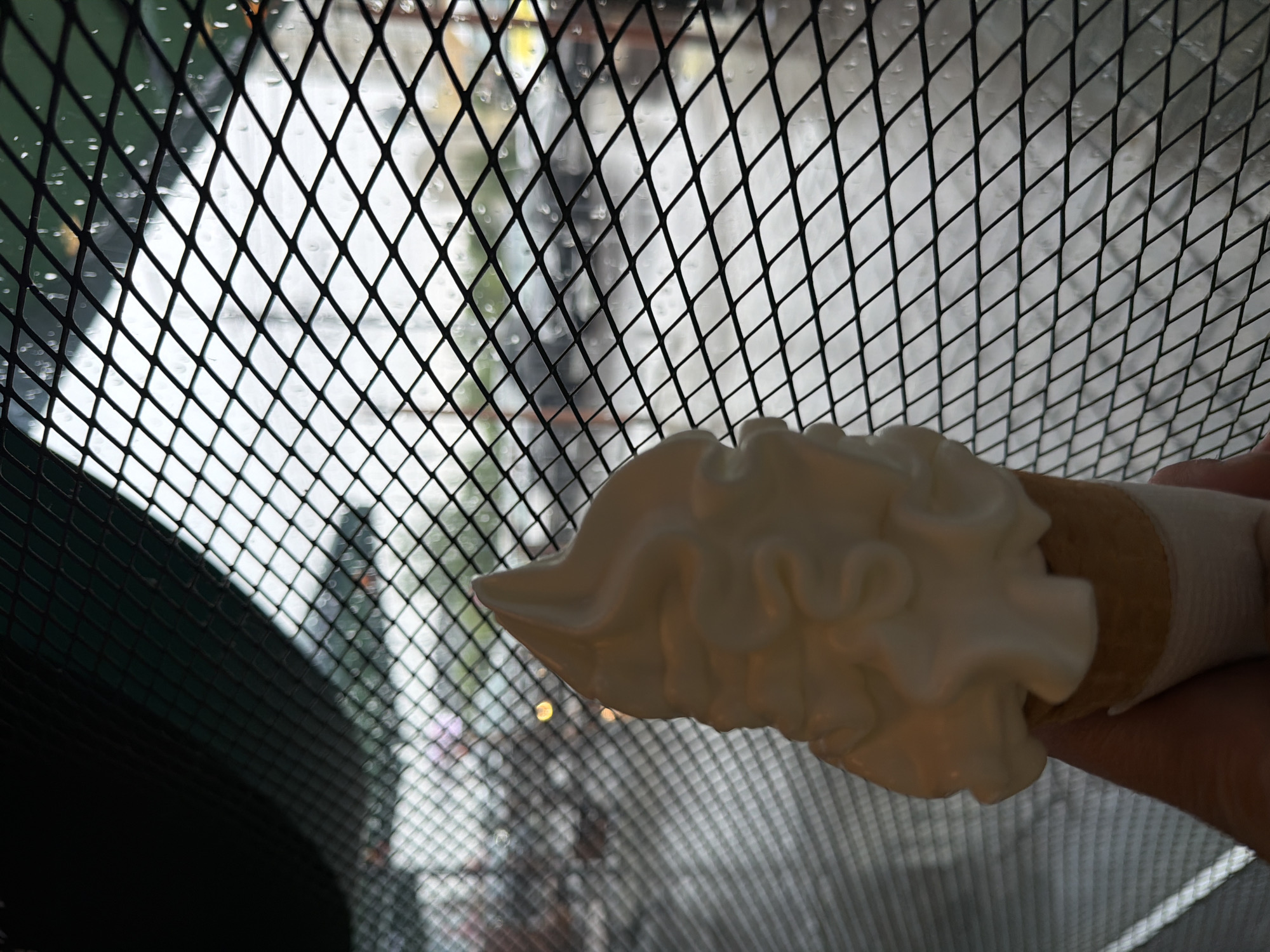 A hand holds an ice cream cone in front of a window with a diamond-patterned metal grate. Through the window, a blurry cityscape is visible. It is Krasnoyarsk, Russia. The cityscape includes tall buildings. The ice cream is white and swirled, sitting atop a tan cone. The person holding the ice cream is wearing a bandage on their hand. The weather appears to be overcast or rainy.