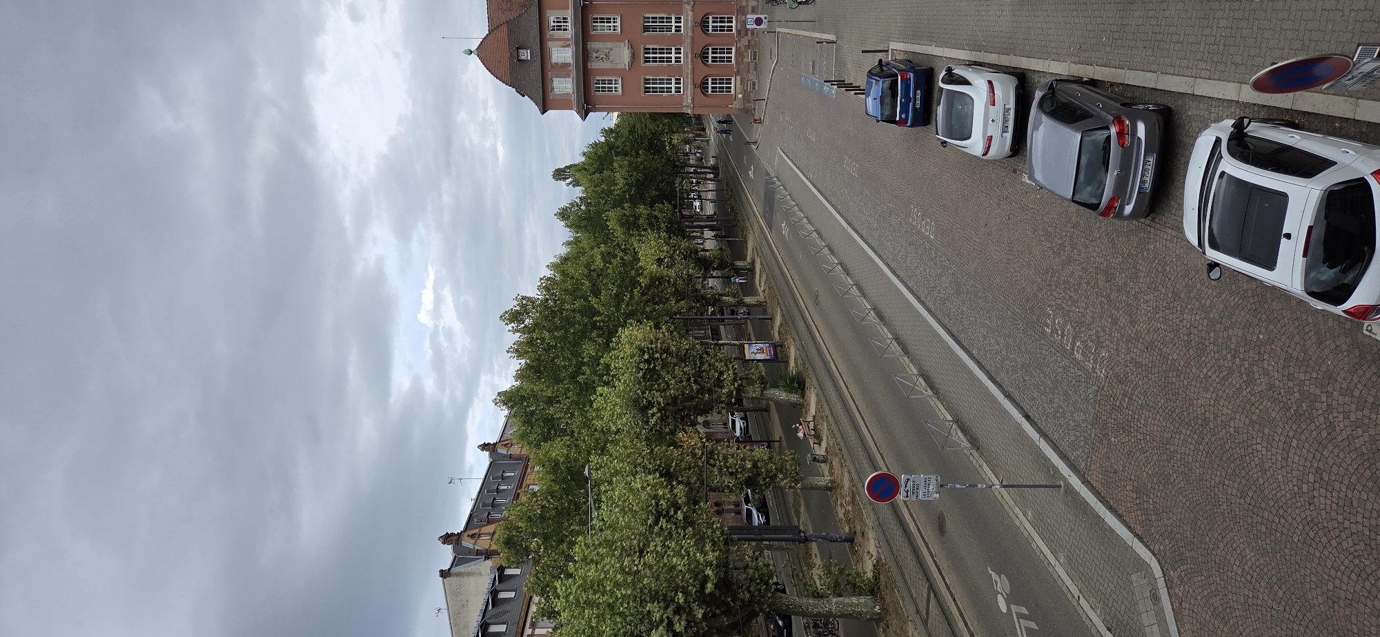 This image depicts a street scene in Strasbourg, France. The perspective is from an elevated viewpoint looking down a tree-lined street with parked cars. The sky is overcast with scattered clouds.
On the left side of the street, there is a row of trees with lush green foliage. Behind the trees, buildings with red-tiled roofs and dormer windows are visible. A streetcar track runs along the cobblestone pavement. A signpost with a red and blue "no parking" symbol and French text is present. Further down the street, a person is sitting on a park bench under the trees, and a trash receptacle is nearby.
The right side of the street features a large building with a salmon-colored facade, white-framed windows, and a red-tiled roof with a green spire. A stone relief is set into the facade above the second story. Several cars are parked along the curb, including a blue hatchback, a white sedan, and a gray hatchback. The street itself is paved with cobblestones. In the foreground, a white hatchback with a sunroof is parked.