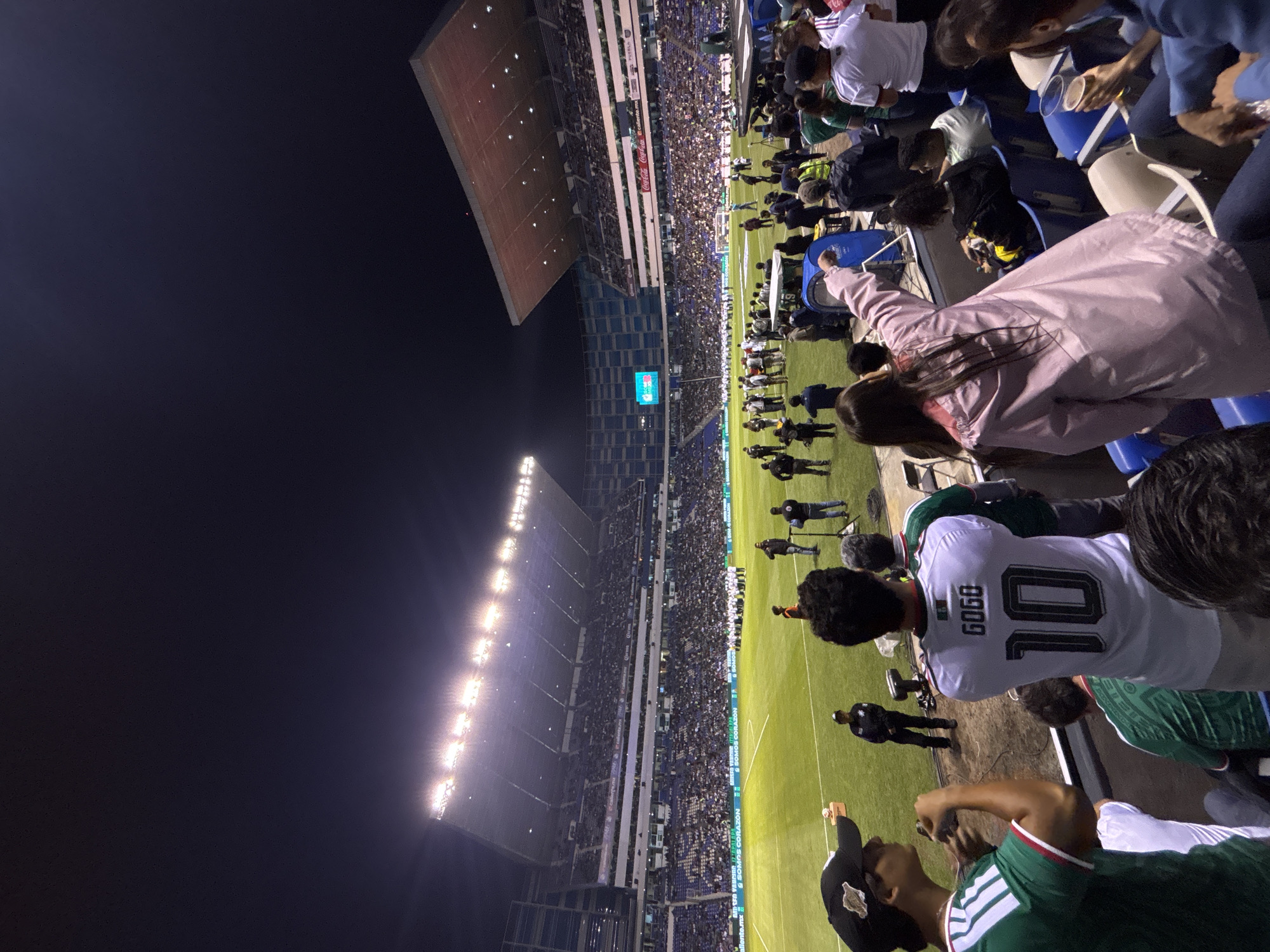 This image captures a night scene at Estadio Cuauhtémoc in Puebla, Mexico, during a soccer game. The stadium is filled with spectators, and the field is brightly lit by stadium lights. People are wearing jerseys, with at least one displaying the number "10" and the name "GOGO". Security personnel are visible along the edge of the field. The stands are packed, and a large section of the stadium is covered by a roof. A scoreboard is visible displaying information about the game.
