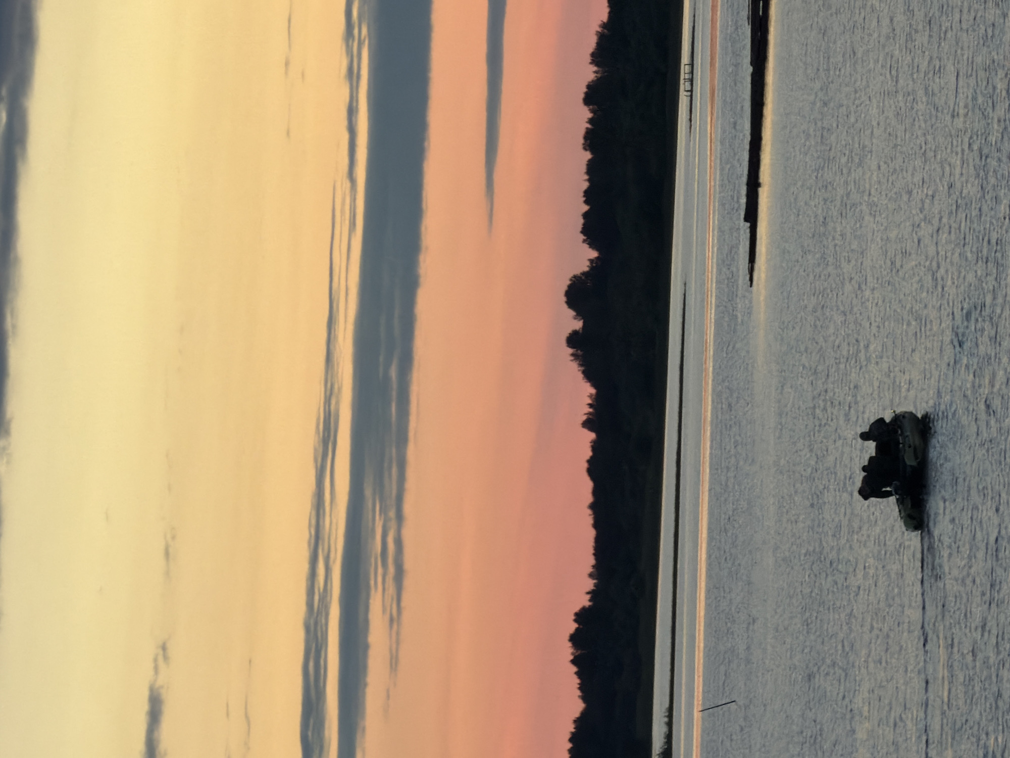 An inflatable boat with three silhouetted figures is on a body of water at dusk. The sky above transitions from yellow at the top to pink and then orange closer to the horizon, with some dark gray clouds. A dark tree line borders the water in the background. The water is rippled and reflects the sky's colors.