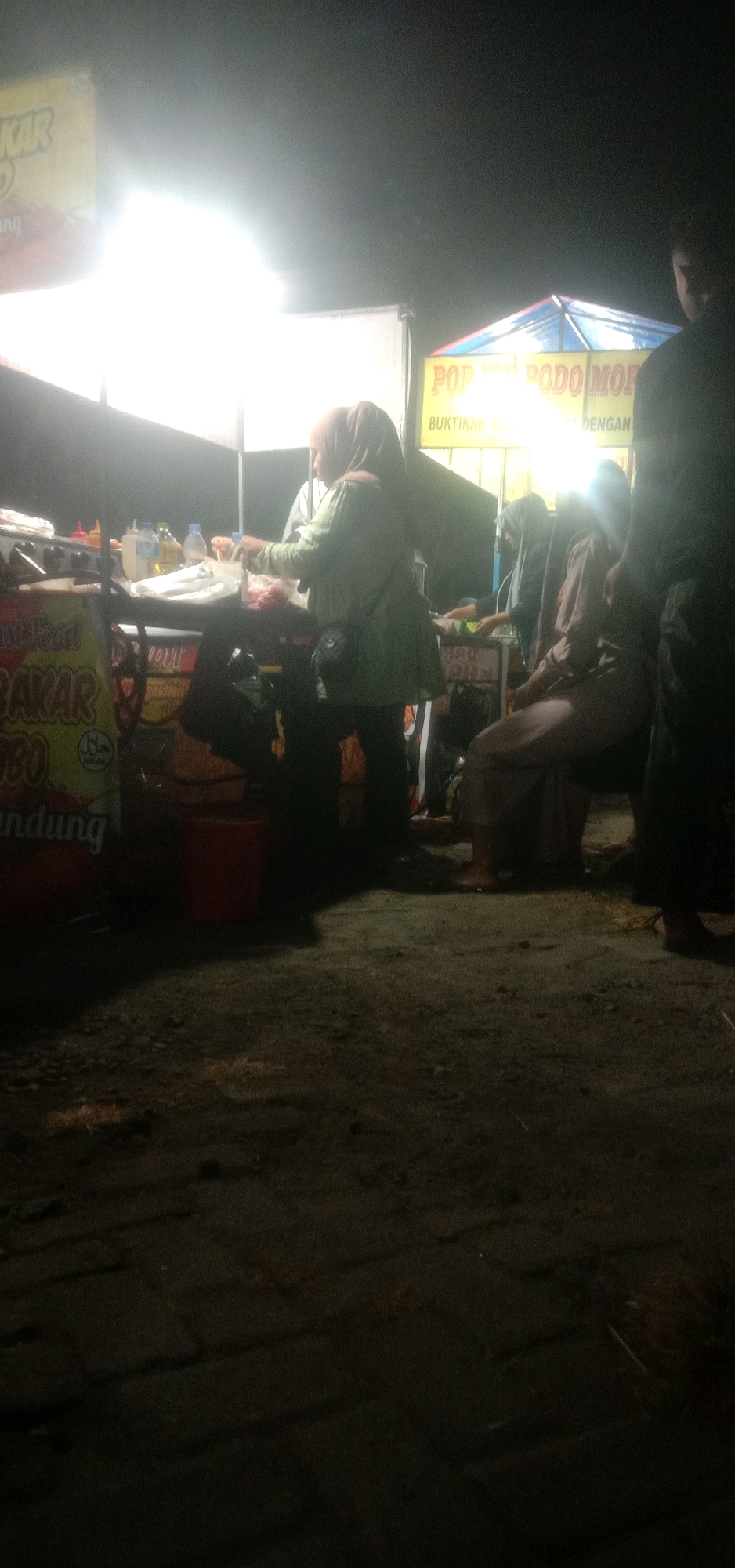 The image captures a nighttime scene at an outdoor food stall in Banjar Dowo, Indonesia. A vendor, a woman wearing a hijab and a green top, is actively preparing food. She stands behind a stall displaying food items and bottles of condiments. To her right, another woman, also wearing a hijab, sits on a stool, observing. Several other individuals are present, their figures mostly obscured by darkness or the bright lighting of the stall. The ground appears to be a dirt or gravel surface, typical of a street or market setting. In the background, a brightly lit sign advertising "POP PODO MORE" is visible, suggesting a food product or service. The overall atmosphere is that of a bustling street food vendor operation at night.