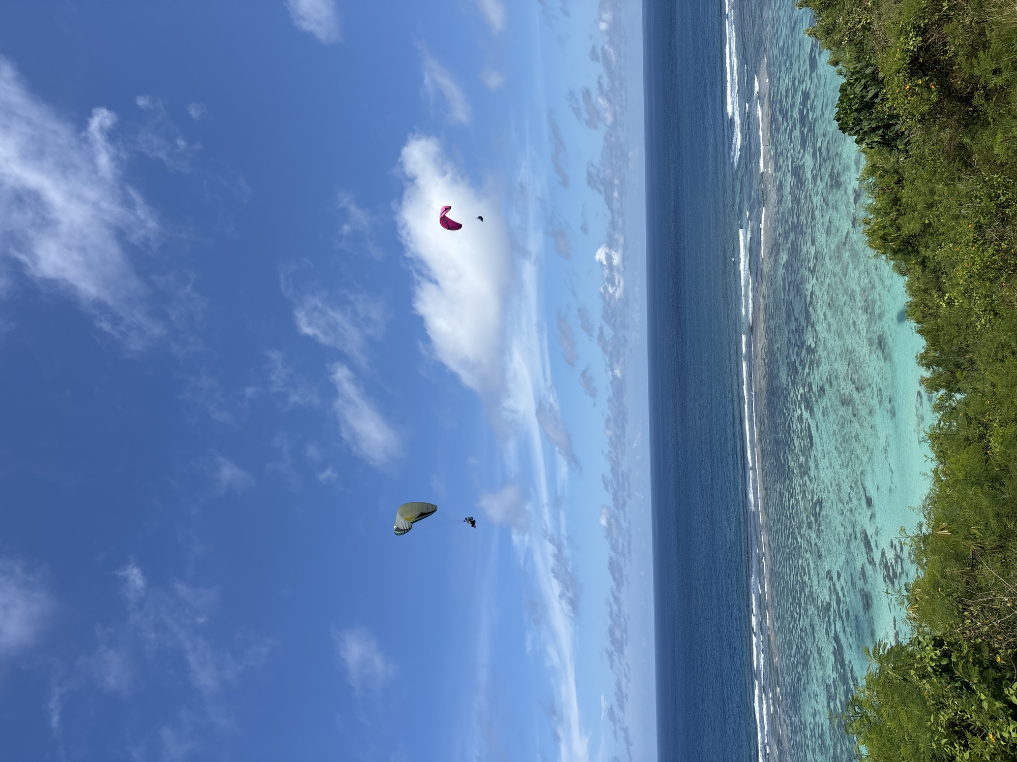 The image shows a coastal scene in Benoa, Indonesia. The sky is blue with scattered white clouds. Two paragliders are visible in the sky, one is red and the other is yellow with a dark colored individual hanging below. The ocean is two-toned, with deep blue water transitioning to lighter turquoise near the shore. Waves are breaking along a coral reef. The right side of the image shows a green lush coastline.