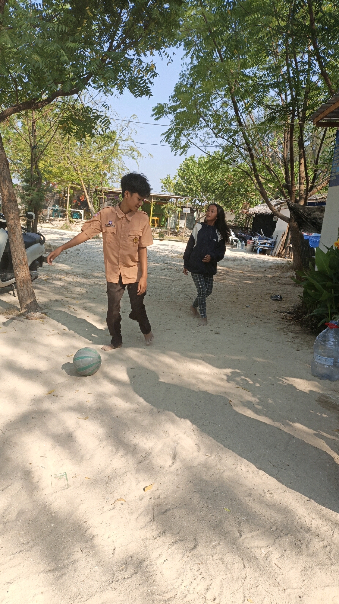 Two individuals, a male and a female, are present in a sandy outdoor area, likely a street or pathway, in Cikal, Indonesia. The male, wearing a tan uniform with badges and dark pants, is positioned on the left, facing the female and looking down towards a green and white ball on the ground. His left arm is extended, and he appears to be in motion, possibly kicking or about to kick the ball. The female, on the right, is wearing a dark jacket with white accents and checkered pants. She is facing the male and looking towards him with a slight smile, her head tilted. She appears to be walking. The environment is characterized by a sandy ground, sparse trees with green foliage casting shadows, and some structures in the background, including what appear to be small buildings and a parked motorcycle. A large plastic bottle of water is visible on the right side of the image. The sky is clear and blue, indicating a sunny day. The overall scene suggests a casual outdoor interaction or play.